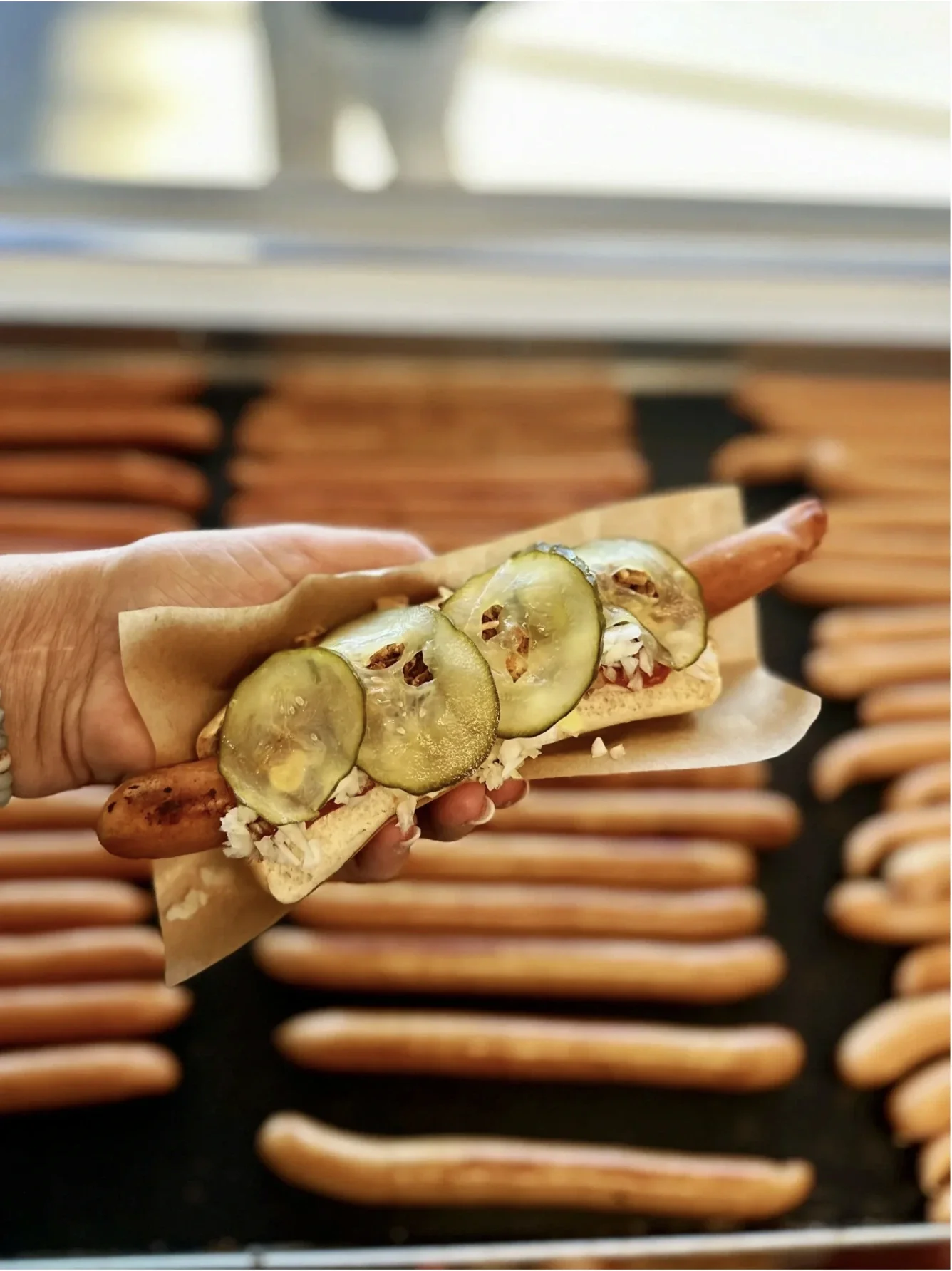En hånd holder en hotdog med sylteagurk, roastbeef, og majssalat, foran et fad med pølser.