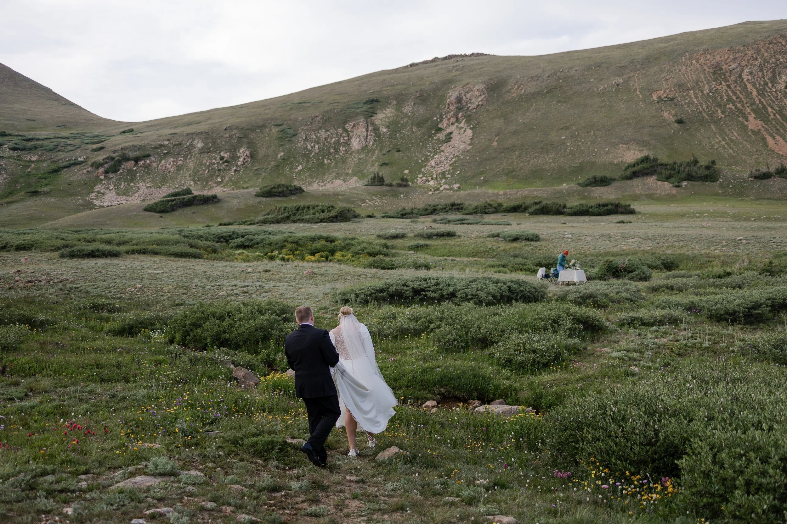 Couple walking in an alpine meadow to approach a picnic setup by their Colorado Elopement Planner in the distance. Photography by Trail Magic Elopements and Micro Weddings.