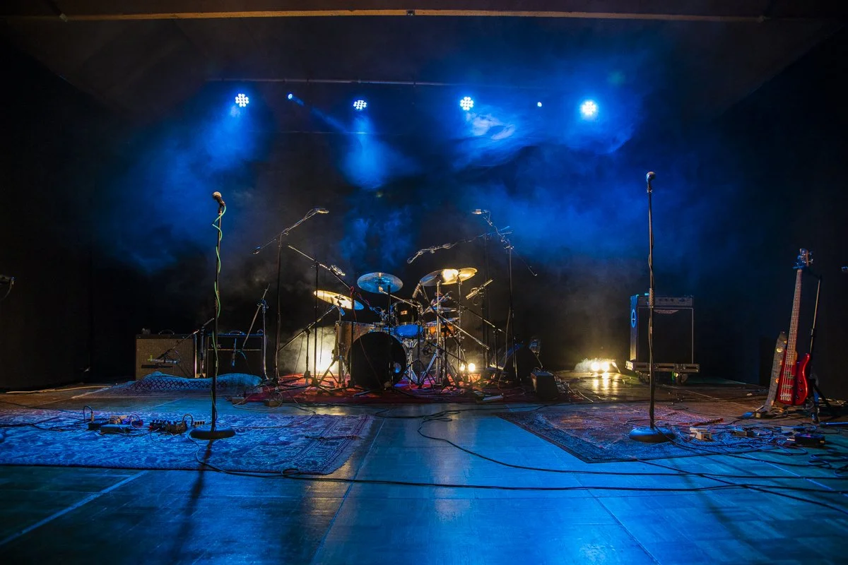 Empty stage with drums, guitars, microphones, and amplifiers illuminated by blue stage lights.
