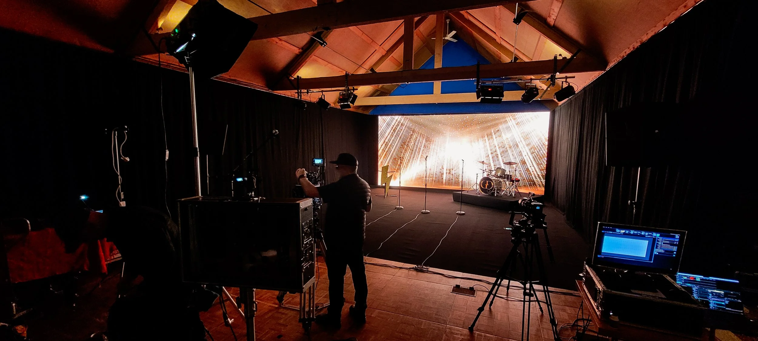 Stage setup with drums and a large digital backdrop displaying radiant, golden beams, in a dimly lit auditorium or studio. Equipment includes cameras, lights, and a person adjusting settings.