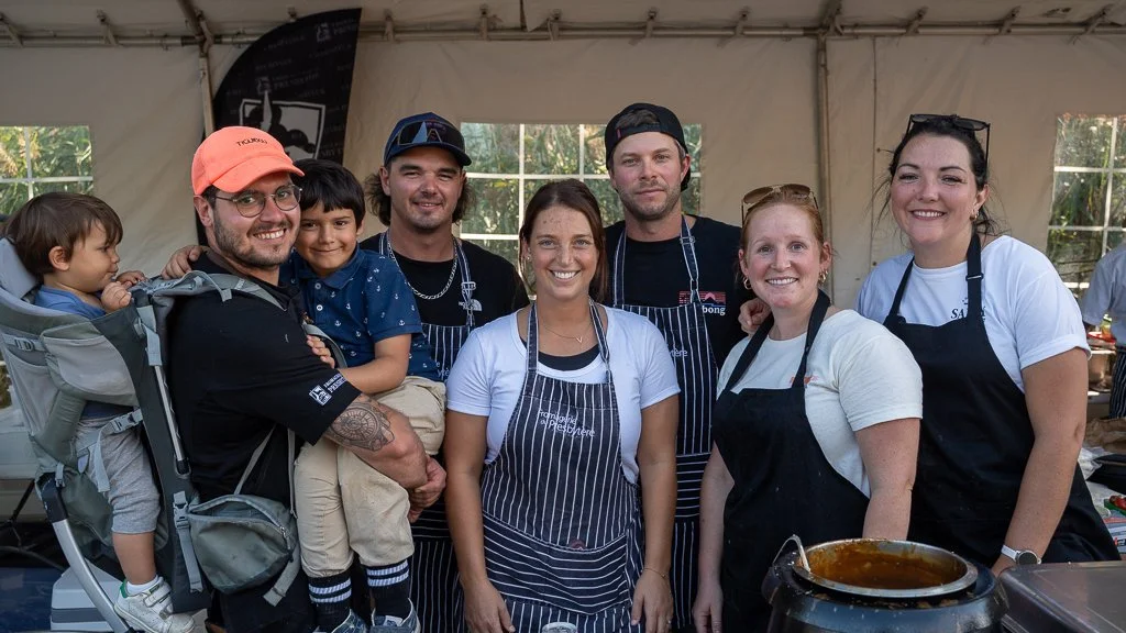 Group of seven people, including children, smiling and posing inside a tent, wearing aprons, possibly at a cooking or barbecue event.