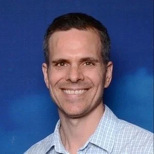 A smiling man with short dark hair, wearing a light blue checkered shirt, against a blue background.