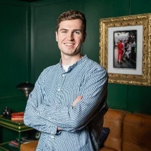 A young man with short hair and a big smile, wearing a blue striped button-up shirt, standing with arms crossed in a room with green walls, a framed picture, and a brown leather couch in the background.