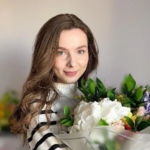 A young woman with long brown hair holding a bouquet of white and pink flowers, smiling slightly, indoors.