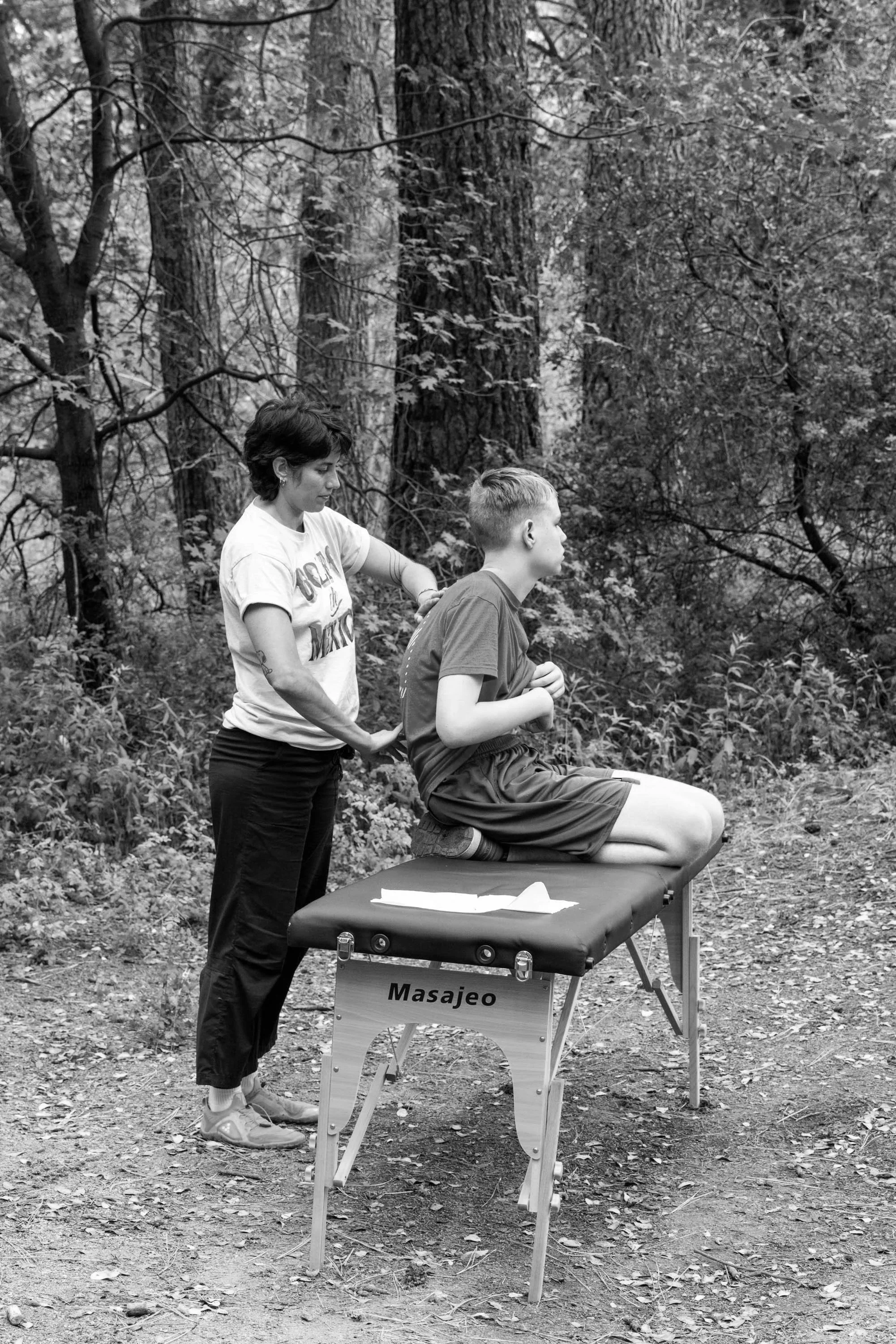 A woman giving a massage to a young man sitting on a massage table in a forest setting.