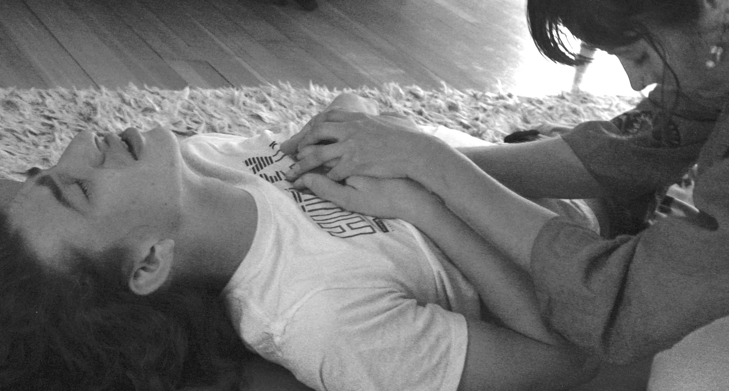 A woman with shoulder-length hair leaning over a woman lying on a carpeted floor, hands clasped on her chest, in an indoor setting.
