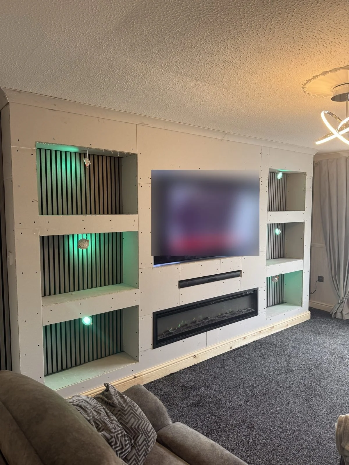 Boarded media wall before plastering, with green-lit slat panel alcoves and electric fire