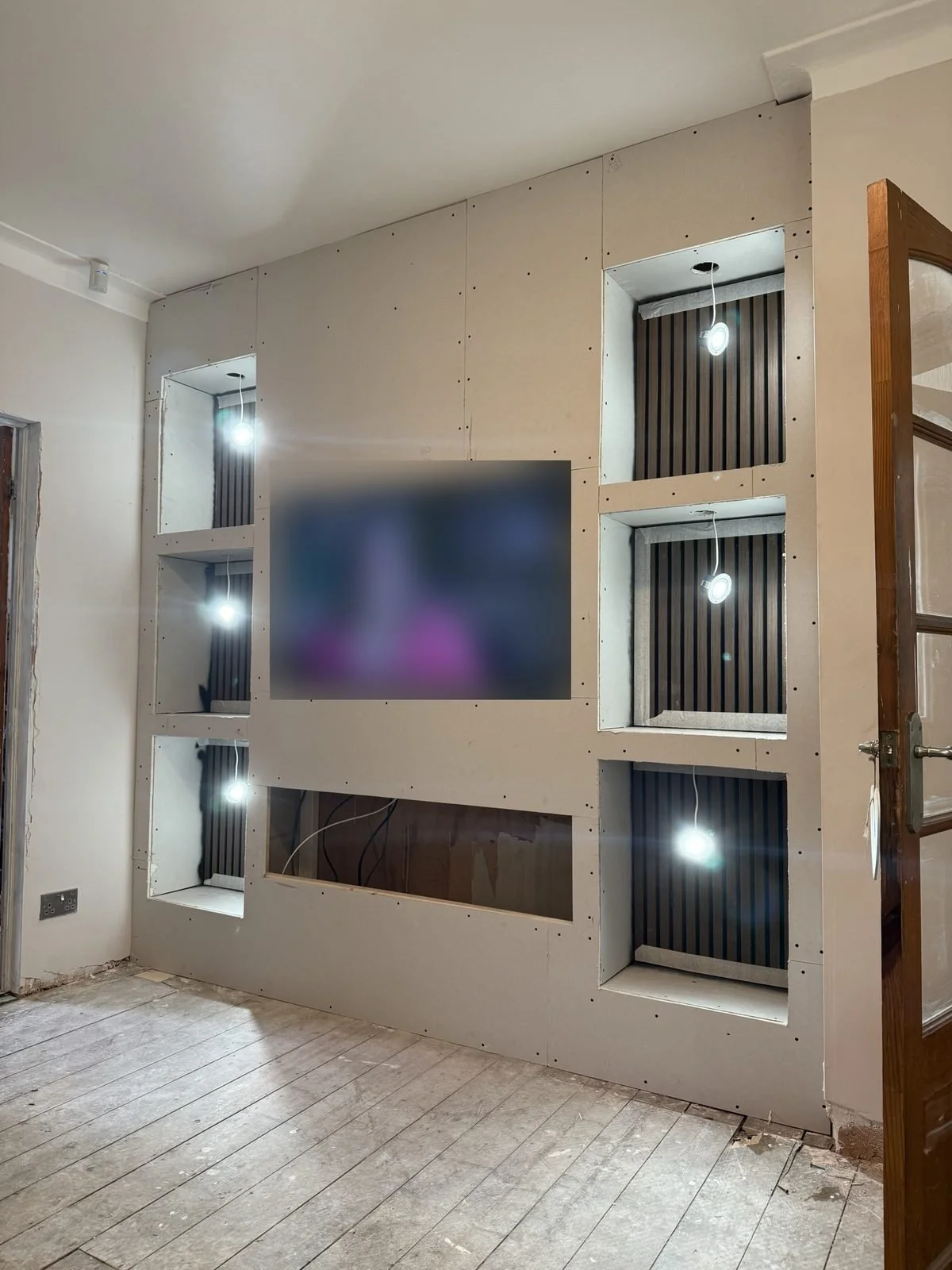 Boarded media wall before plastering, with recessed TV, lit slat panel alcoves and fire opening