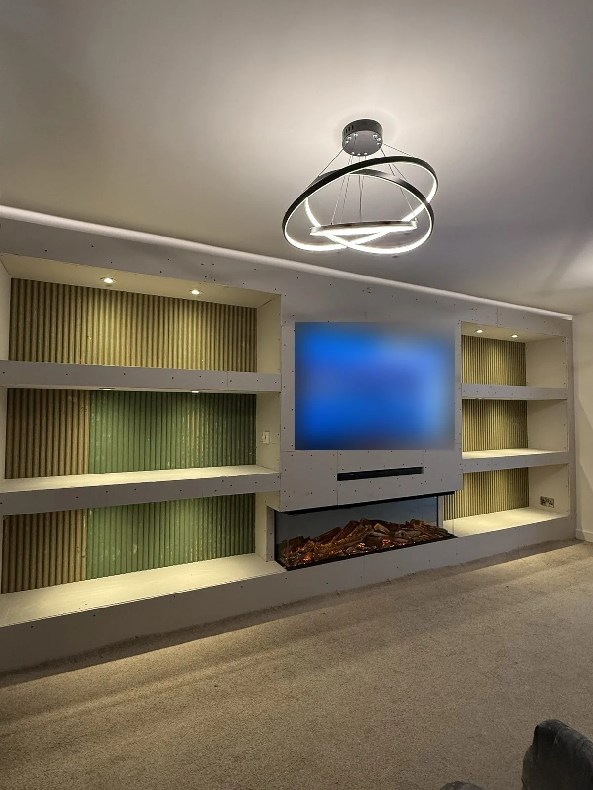 Wide media wall build before plastering, with LED-lit shelves, green slat panel alcoves, TV and electric fire