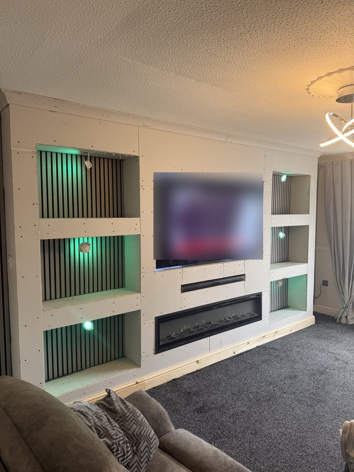 Boarded media wall before plastering, with green-lit slat panel alcoves and electric fire