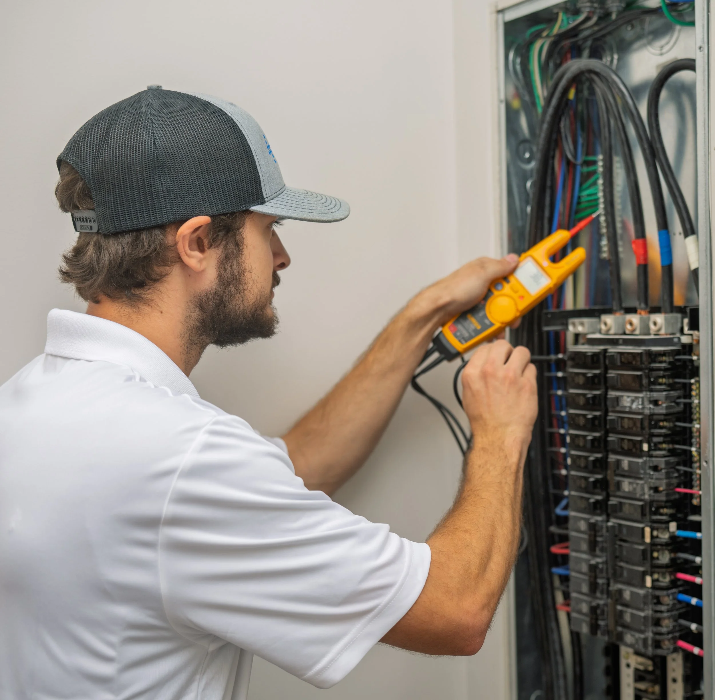 A field engineer wearing a yellow safety helmet and a white t-shirt with 'FE' and 'Field Engineer' printed on it works on electrical outlets on a wall, with cords plugged in and routed along the baseboard.