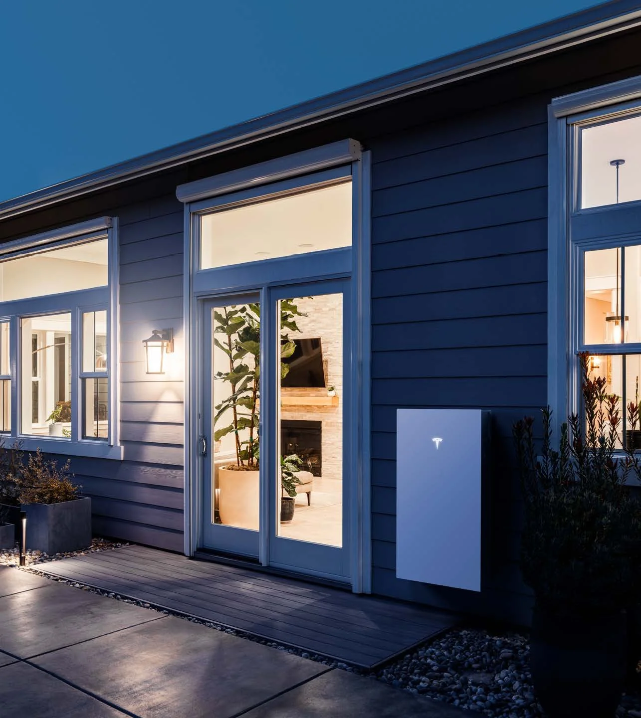 Exterior view of a modern house with blue siding and large windows illuminated from inside, evening setting.