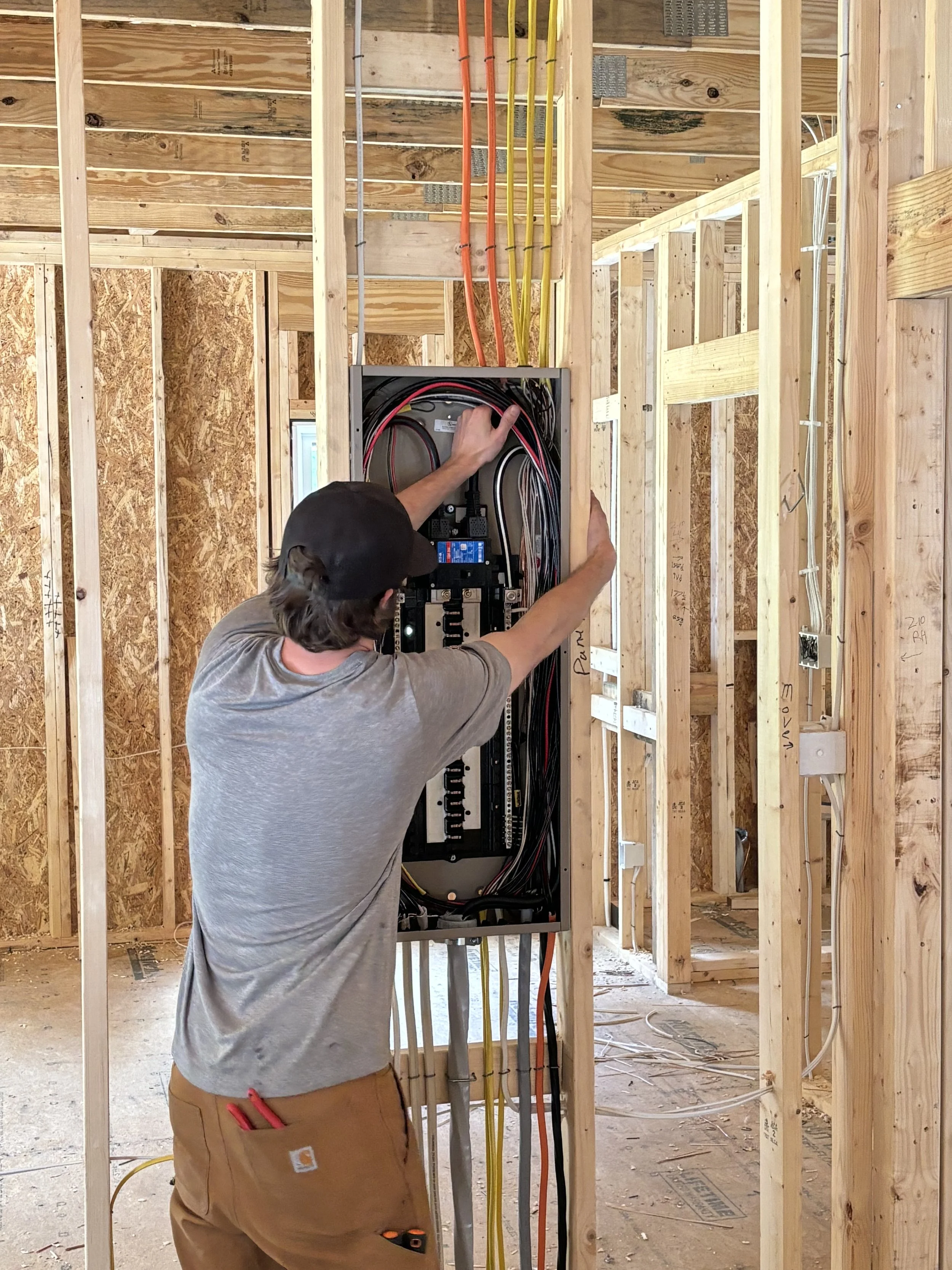 A field engineer wearing a yellow safety helmet and a white t-shirt with 'FE' and 'Field Engineer' printed on it works on electrical outlets on a wall, with cords plugged in and routed along the baseboard.