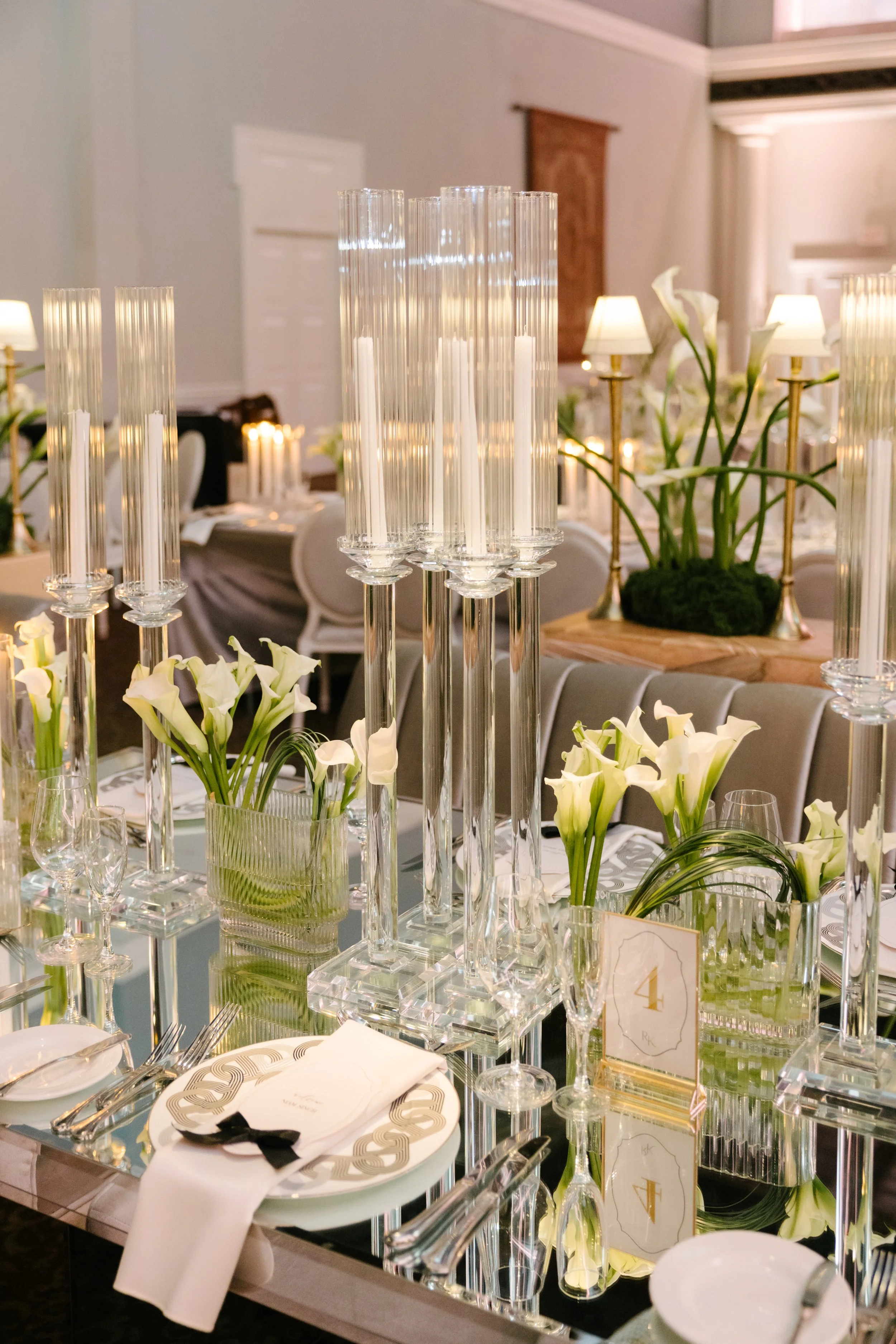 Elegant dining table with tall glass candle holders, white calla lilies in glass vases, gold table numbers, and neatly arranged silverware, set in a fancy event space.