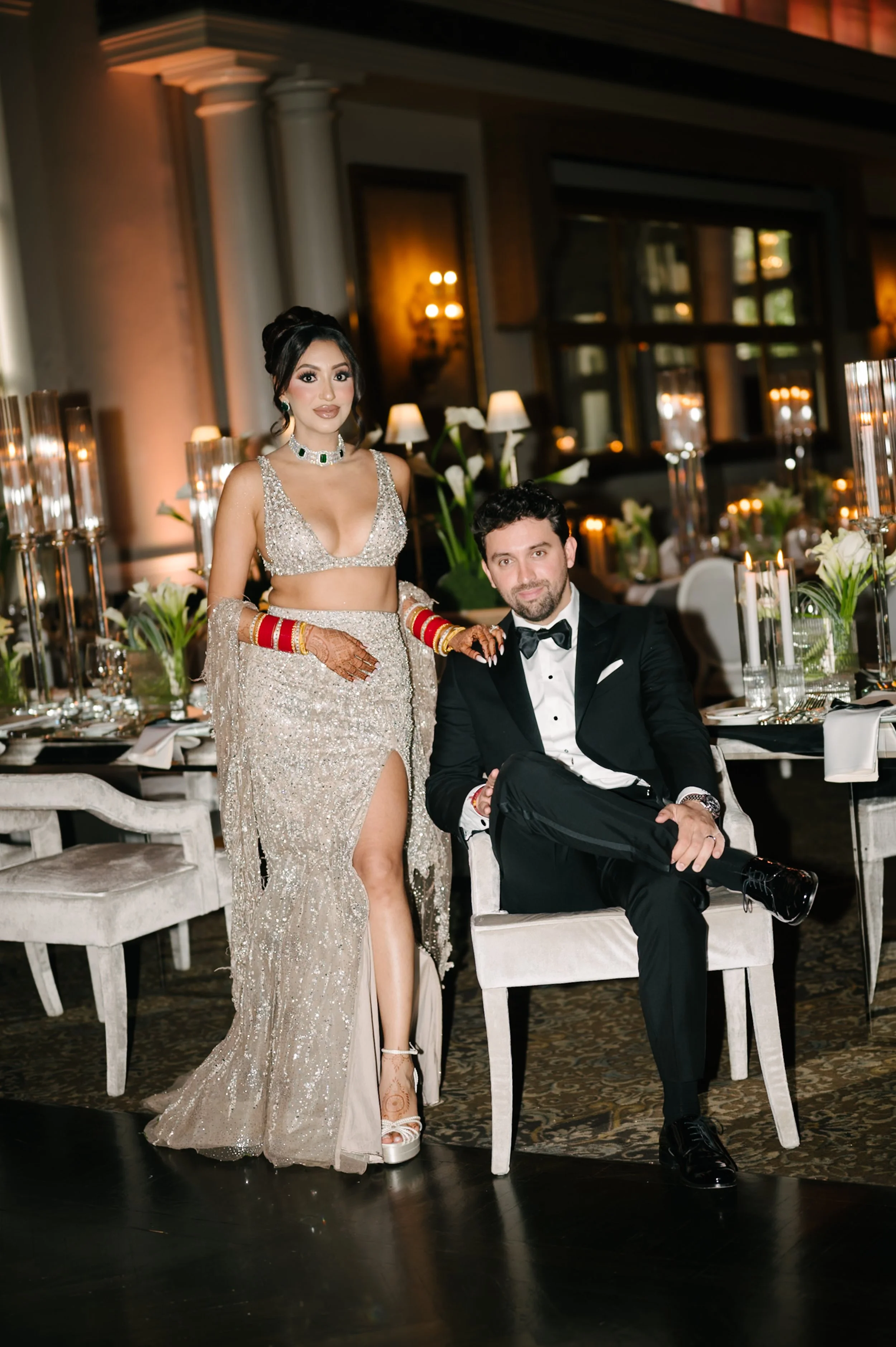 A woman in a silver gown and a man in a tuxedo pose at a formal dinner event, with a decorated banquet table featuring candles and flowers in the background.
