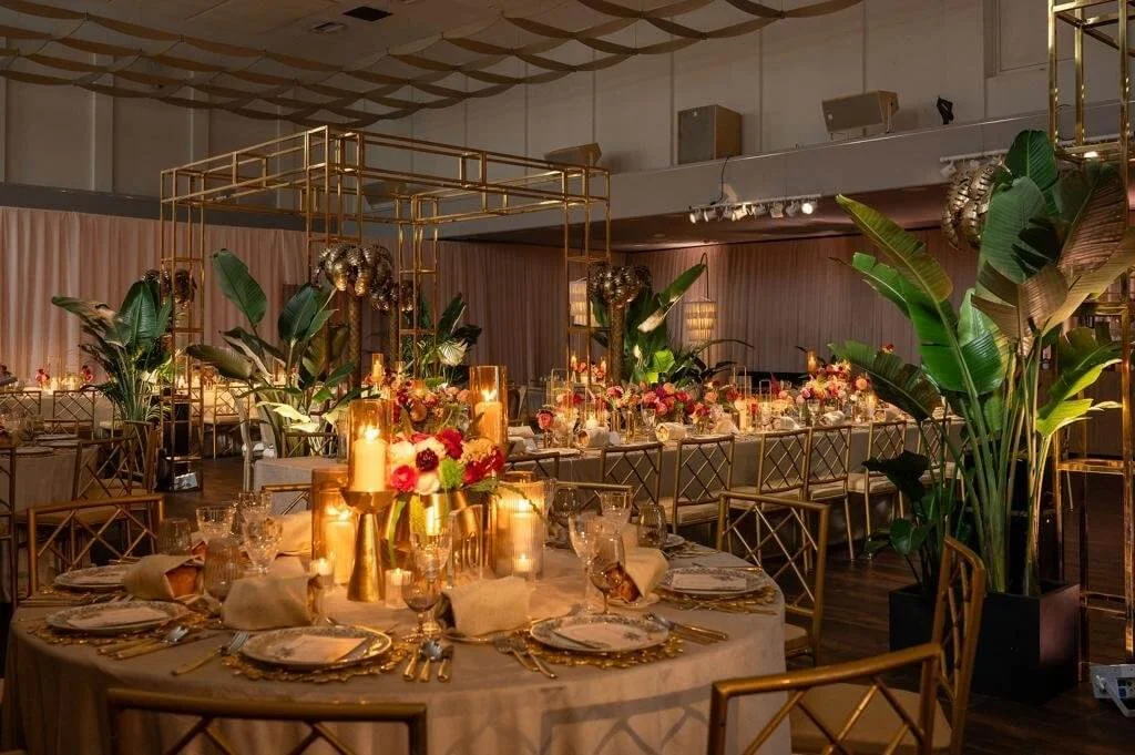 Beautifully decorated event space with round tables set for a formal dinner, featuring gold accents, floral centerpieces, candles, and lush green plants.