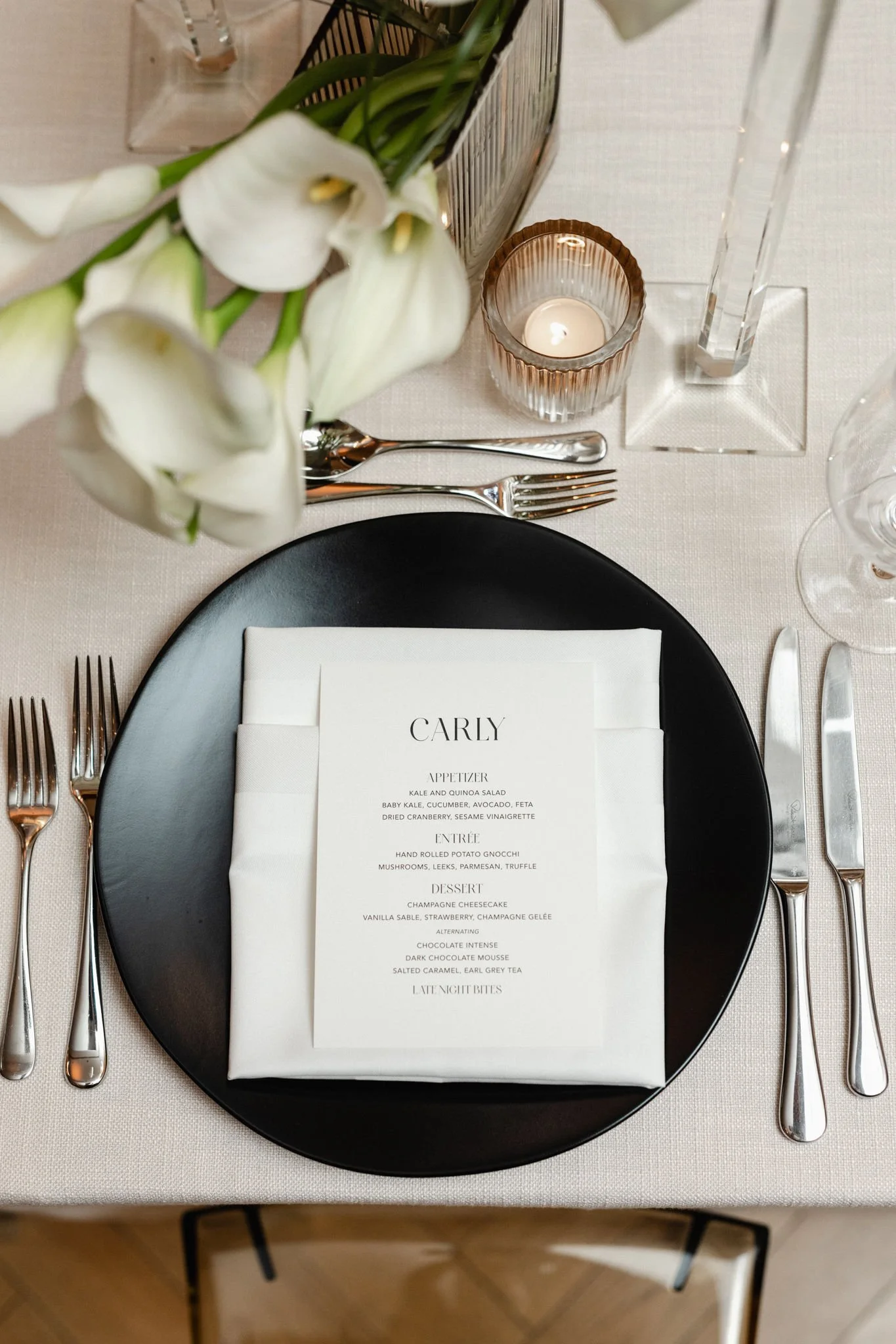 Elegant table setting with a black plate, white napkin, and a menu card labeled "Carly." Silverware arranged on both sides of the plate, a small candle in a glass holder, and floral arrangements in a clear vase on a cream-colored tablecloth.