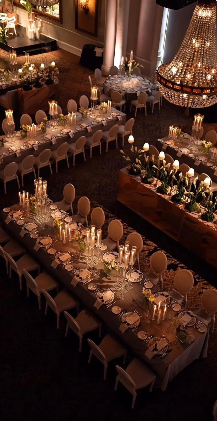 Elegant banquet hall with long dining tables set with white plates, napkins, glassware, candles, and floral centerpieces, illuminated by a large chandelier.