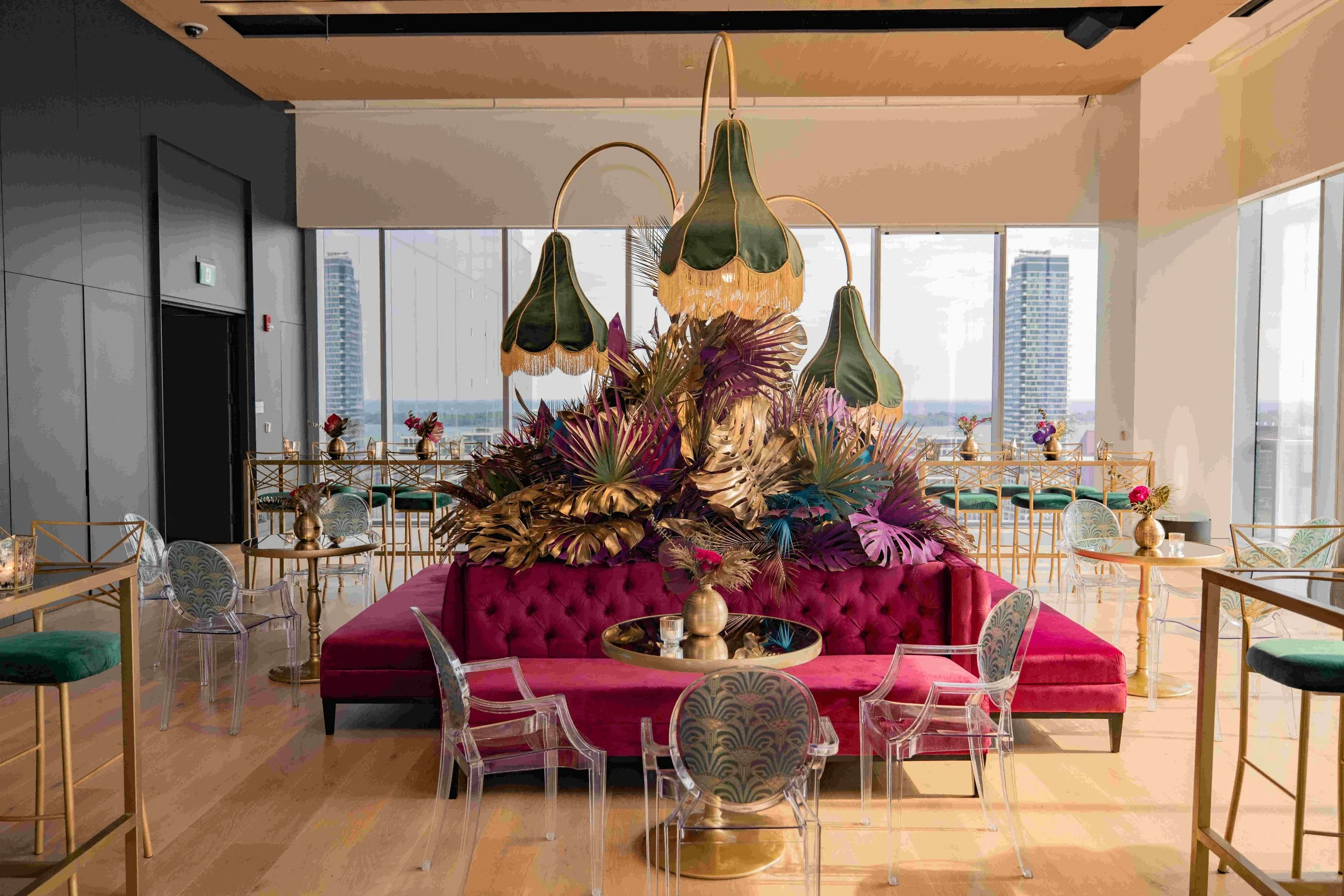 Interior view of a modern room with a large floral arrangement in the center, including oversized leaves and plants, topped by green lampshades with gold fringe, surrounded by colorful chairs and tables with flowers, in a high-rise building with large windows and city skyline view.
