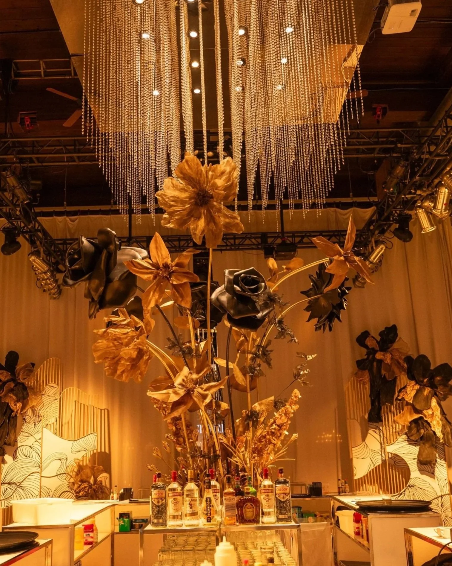 Elegant bar setup with a large floral arrangement featuring black and gold flowers, illuminated by decorative golden lighting, with bottles of alcohol on display and a dramatic ceiling with hanging crystal strands.