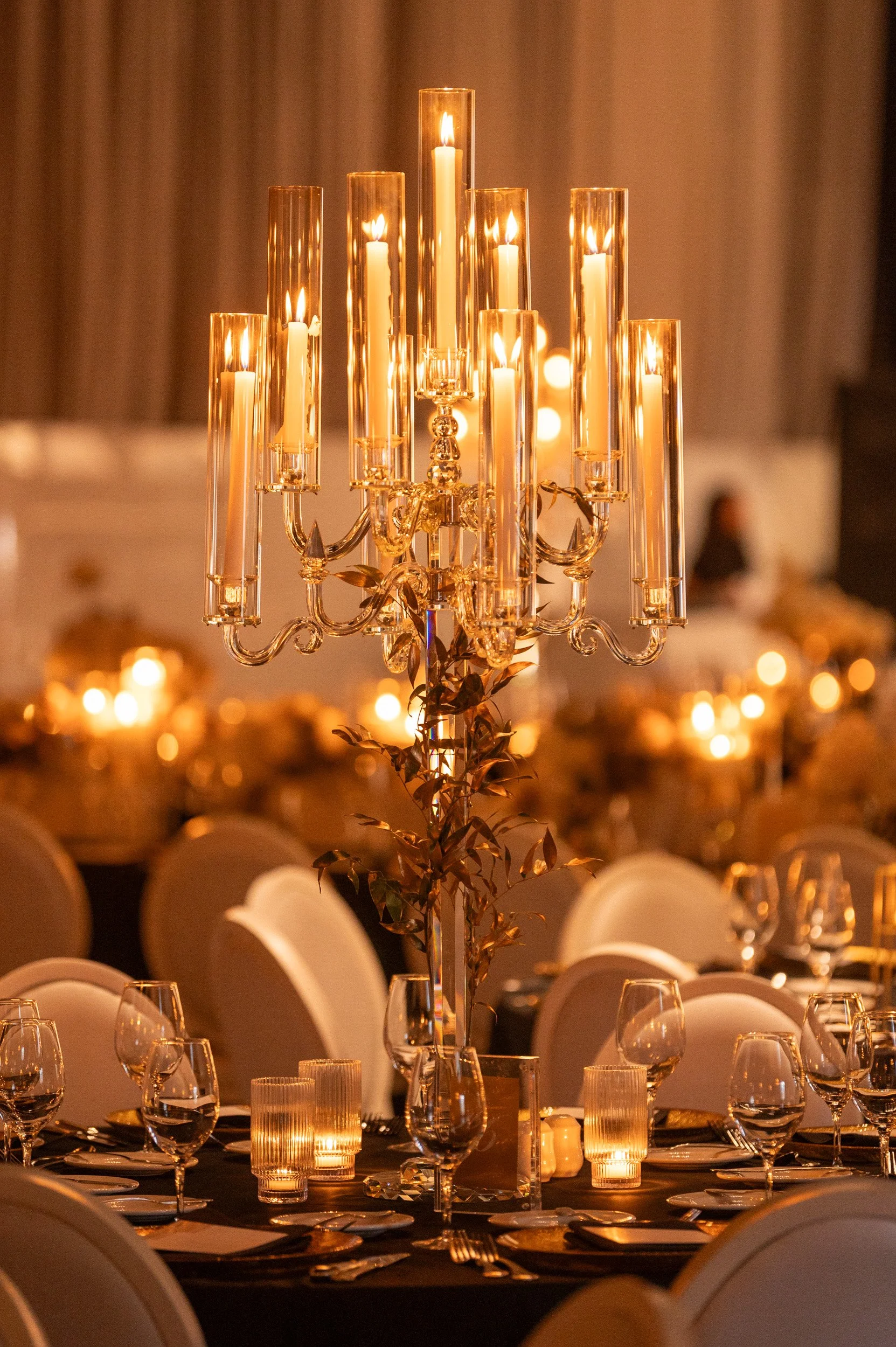 Elegant banquet table decorated with a tall, gold candelabra holding lit candles, surrounded by glasses, silverware, and white chairs, with warm ambient lighting.