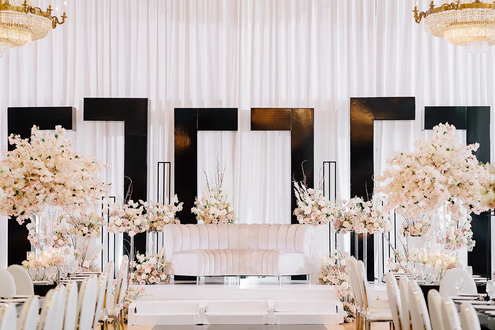 Elegant event stage with white drapery, floral arrangements, black letters, and a pink velvet sofa.