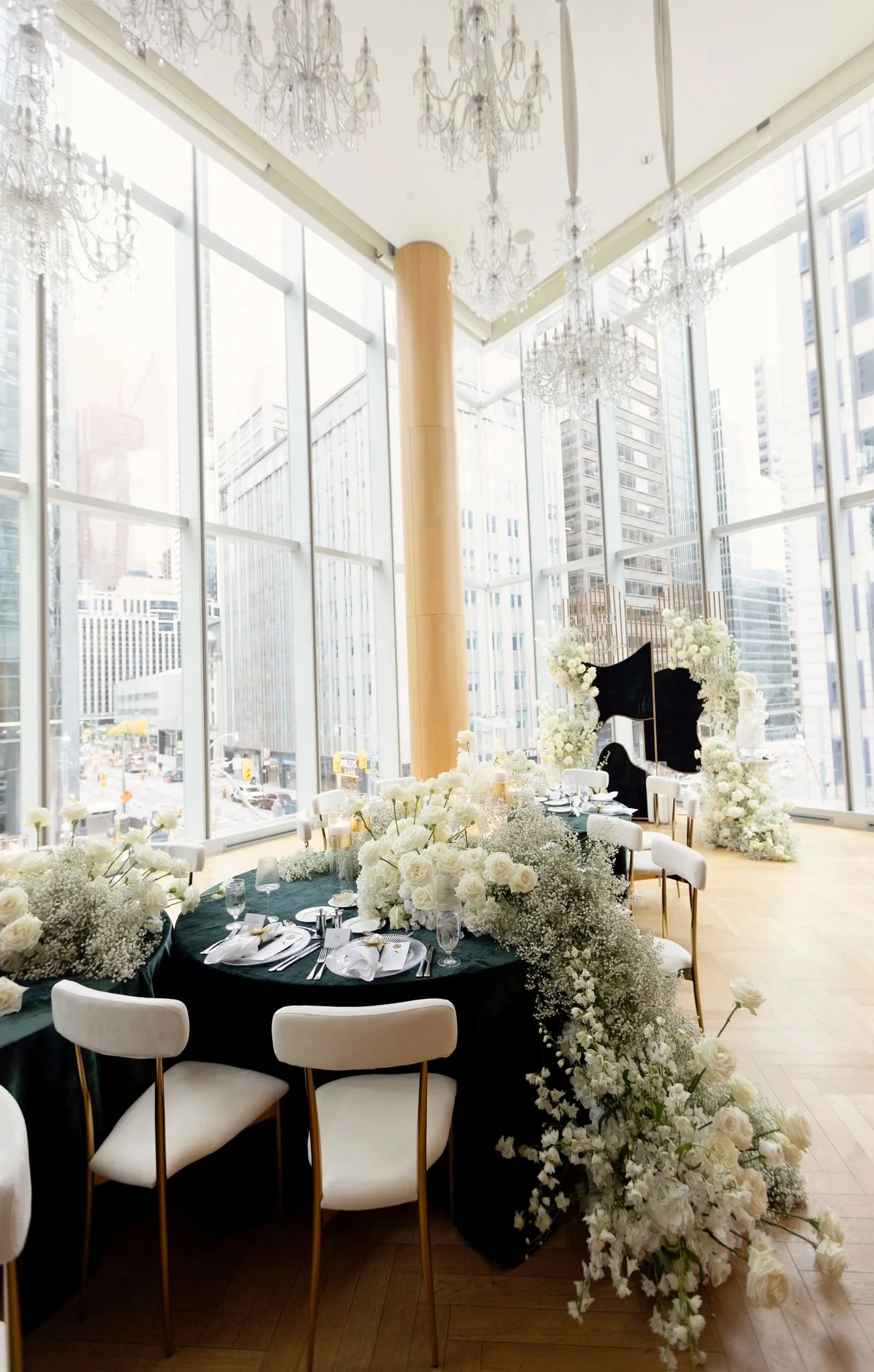 Elegant indoor wedding reception setup with round table, white floral arrangements, and a modern backdrop in a high-rise venue with large windows overlooking city buildings.