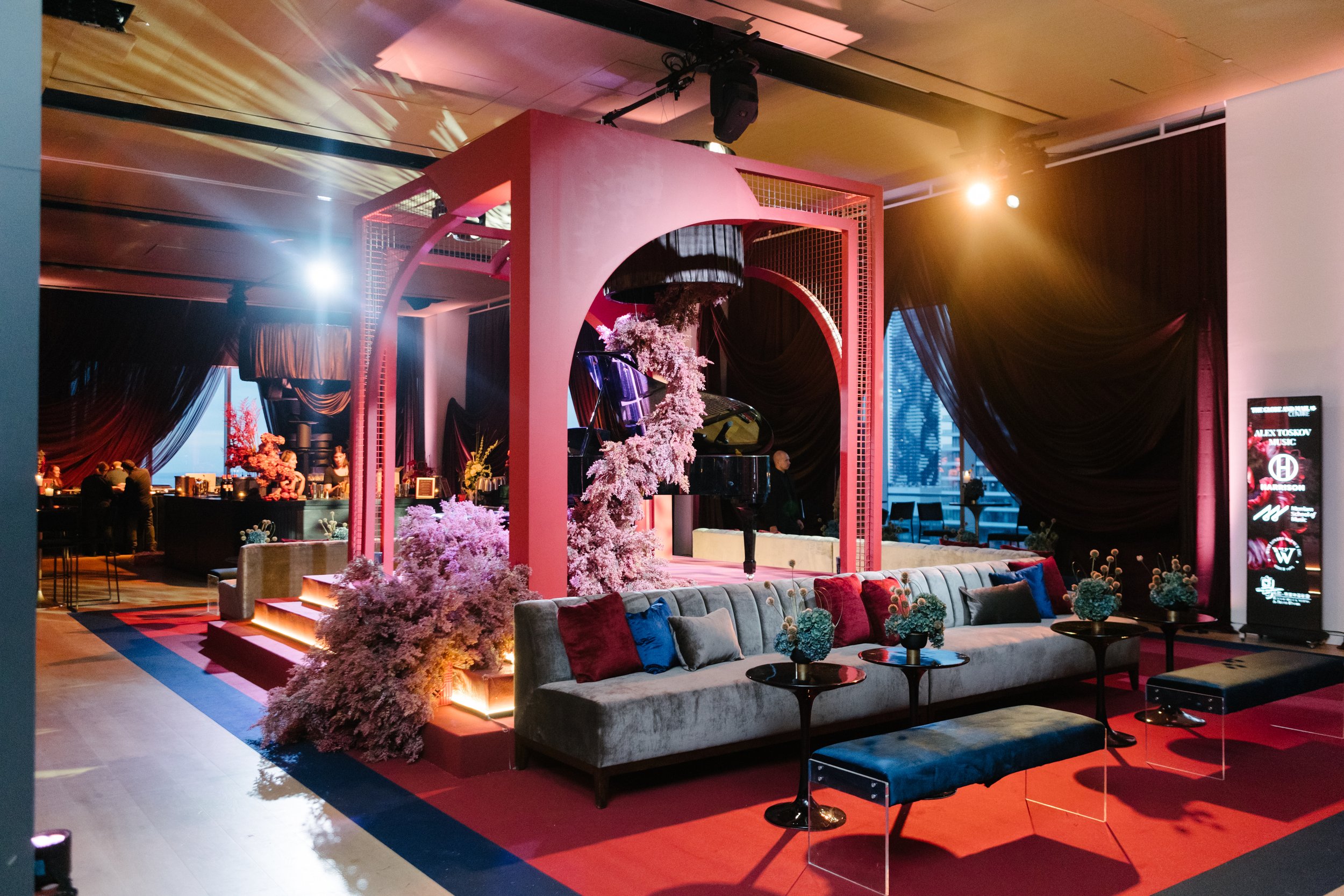 An elegant event space with a grand piano surrounded by pink and white floral arrangements, layered rugs, a long sofa with colorful pillows, small black tables with flower arrangements, and dark curtains with city views in the background.