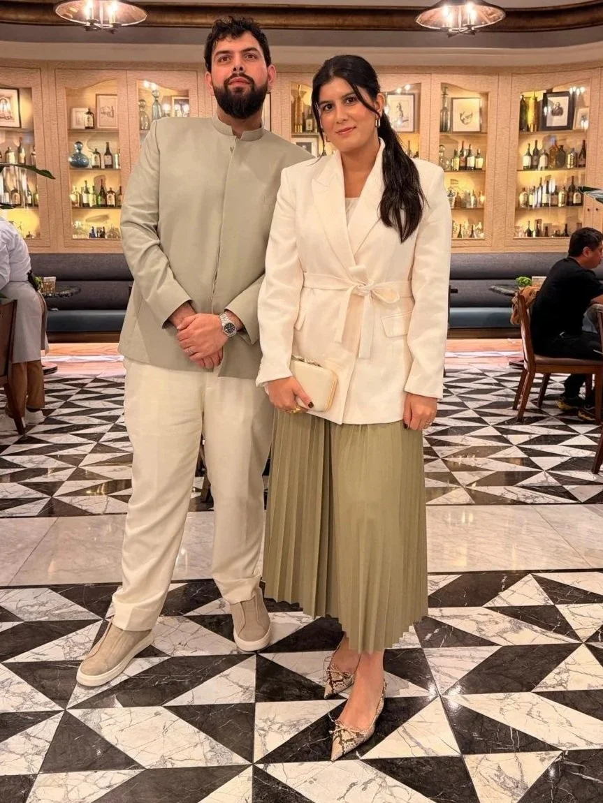A man and woman posing together in a dining area with shelves of bottles and decorative items behind them. The man is wearing a beige suit with a mandarin collar, and the woman is dressed in a white blazer, beige pleated skirt, and snakeskin heels, holding a clutch.
