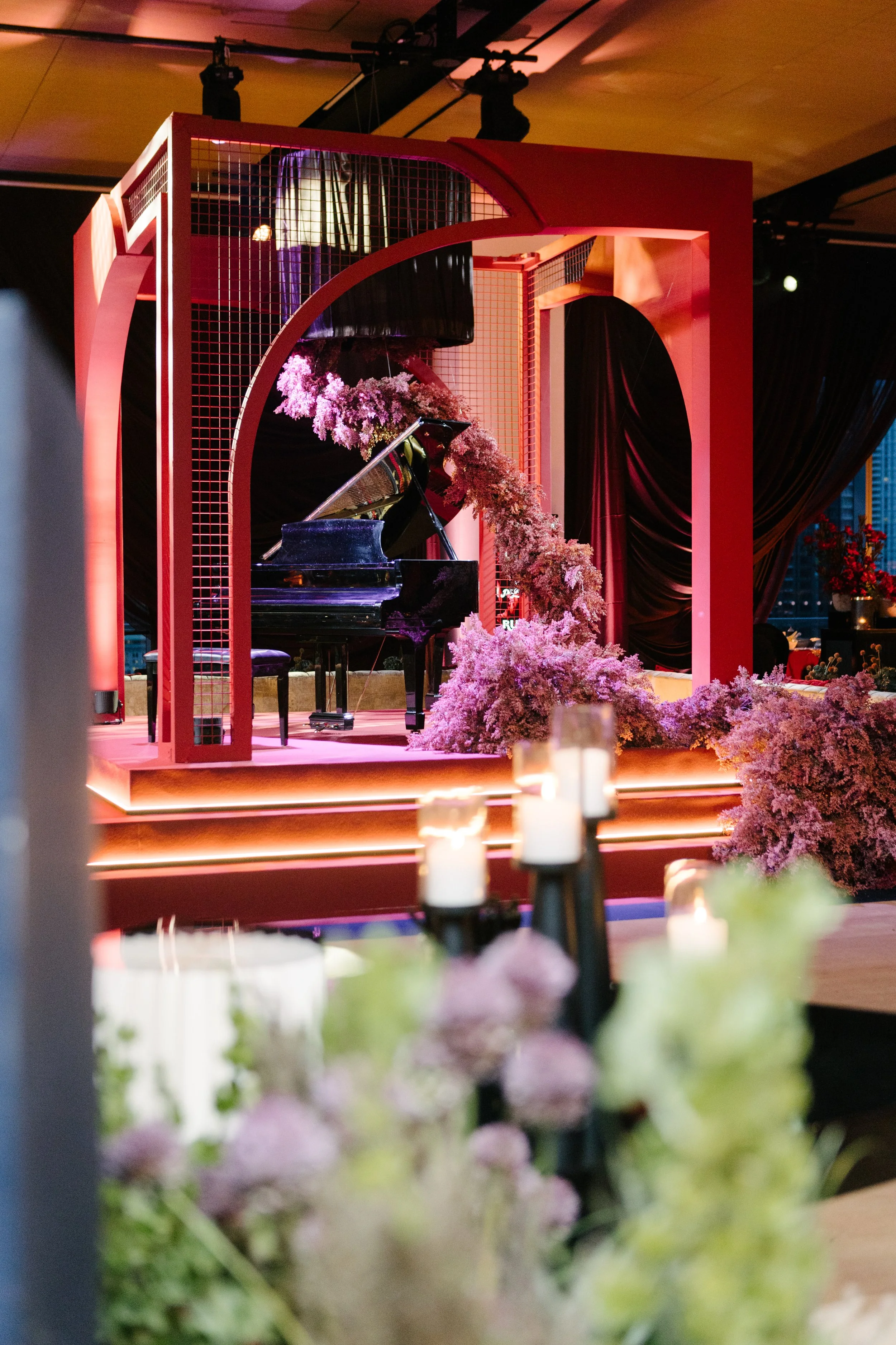 A stage with a black grand piano surrounded by pink floral arrangements and pink lighting, set for a performance or event.