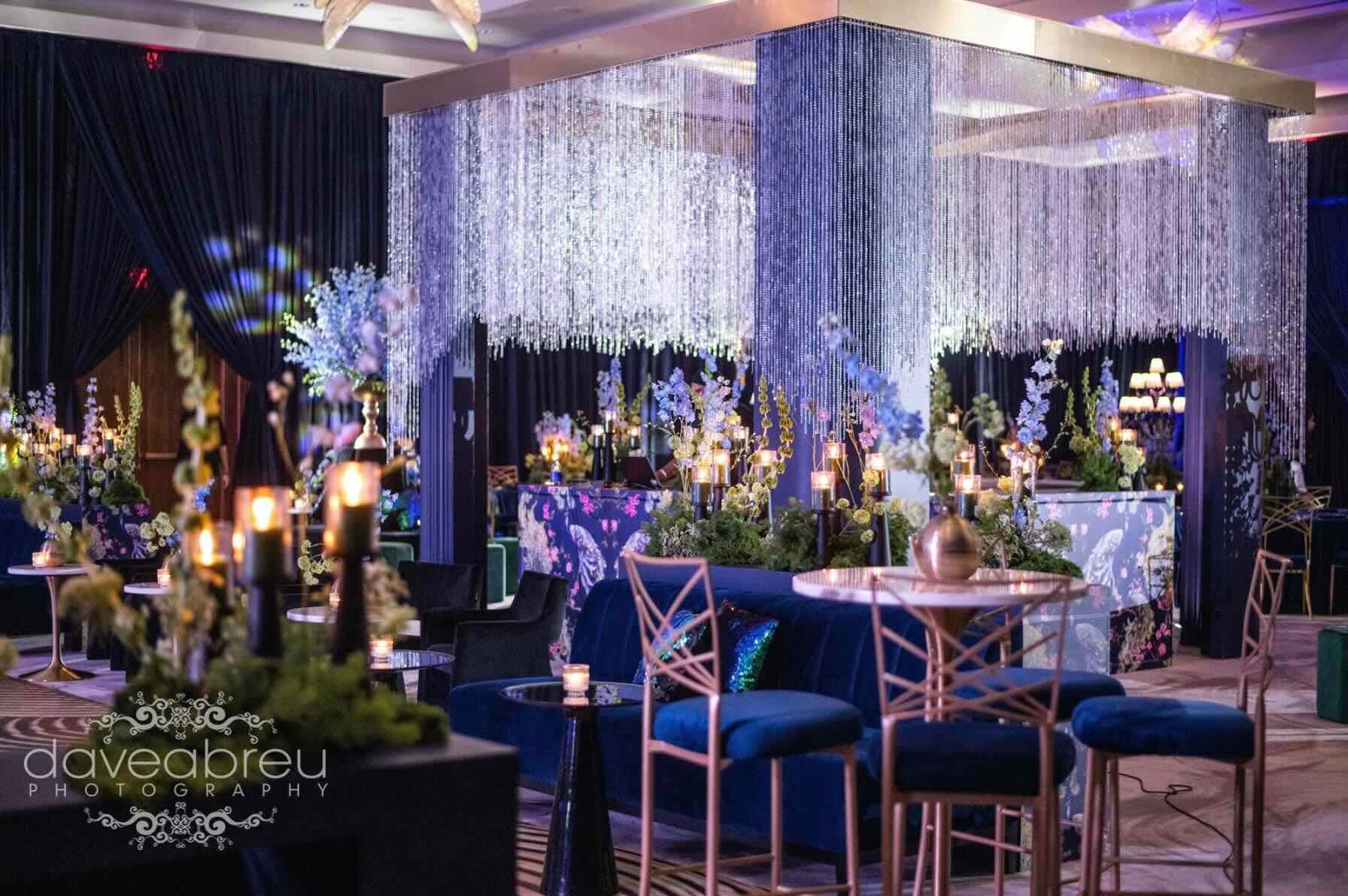 Elegant event space with colorful lighting, floral arrangements, candles, velvet seating, and a decorative curtain of hanging crystals or beads.