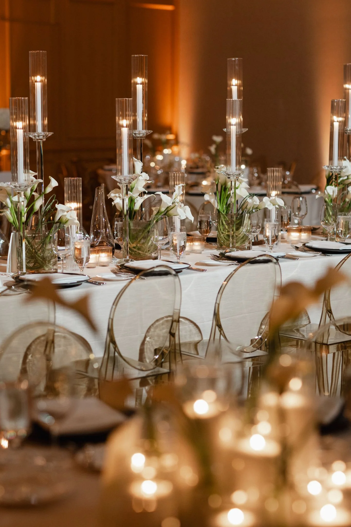Elegant banquet table decorated with tall glass candlesticks, vases of white calla lilies, and candlelight in a warmly lit room.