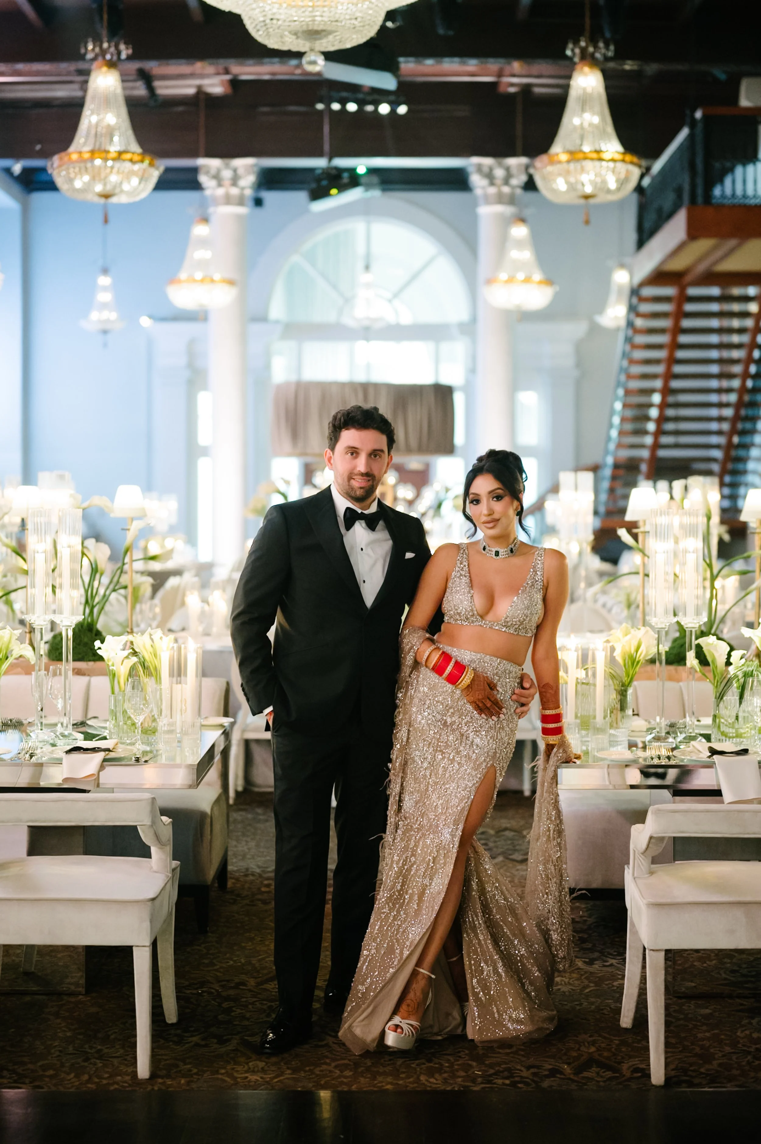 A man in a tuxedo and a woman in a gold, sparkling dress stand together in an elegant, well-lit dining hall decorated with white flowers, candles, and chandeliers.