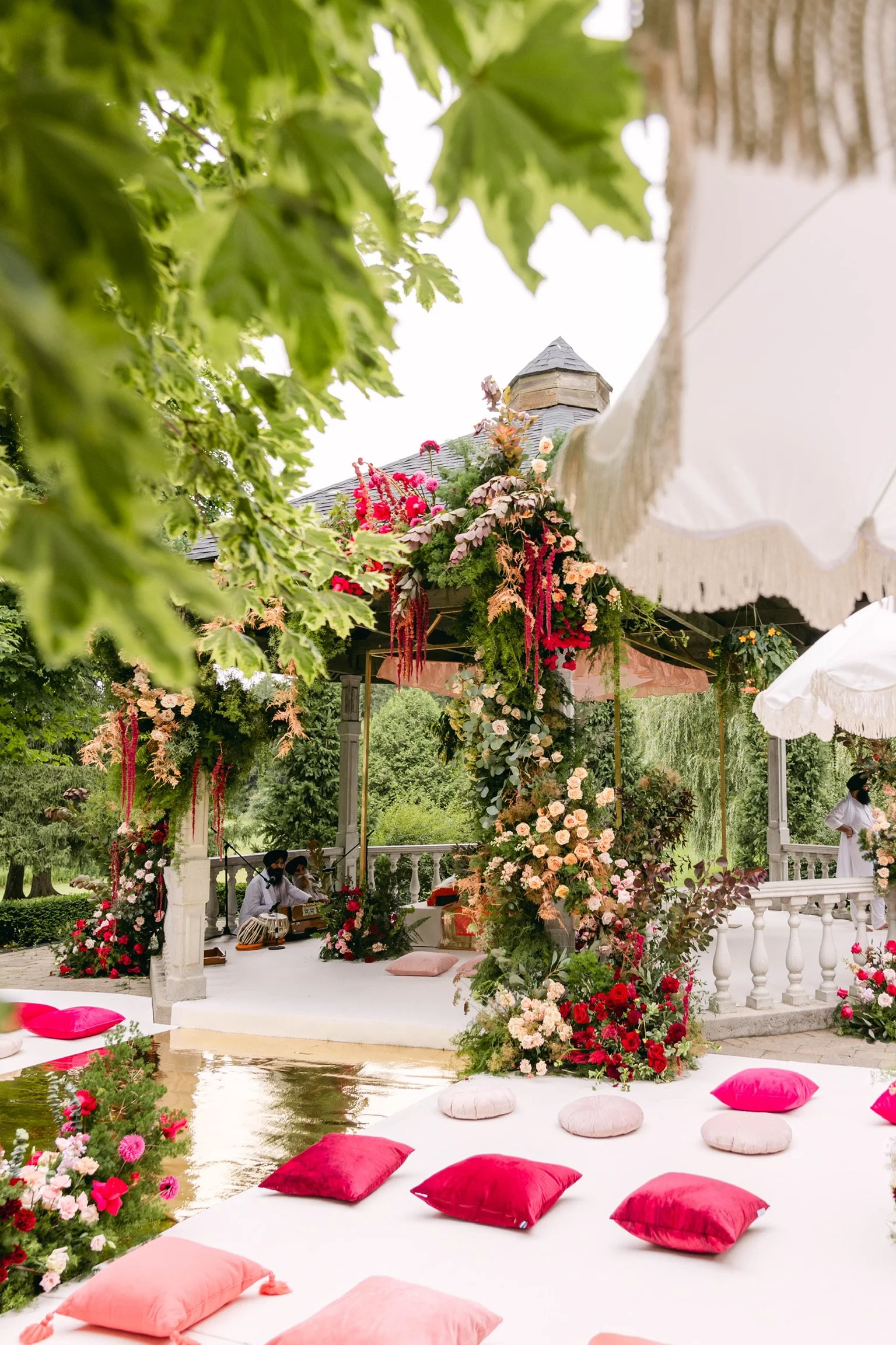 Outdoor wedding or celebration setup with floral arch, cushions, and musicians playing instruments under a canopy, surrounded by trees.