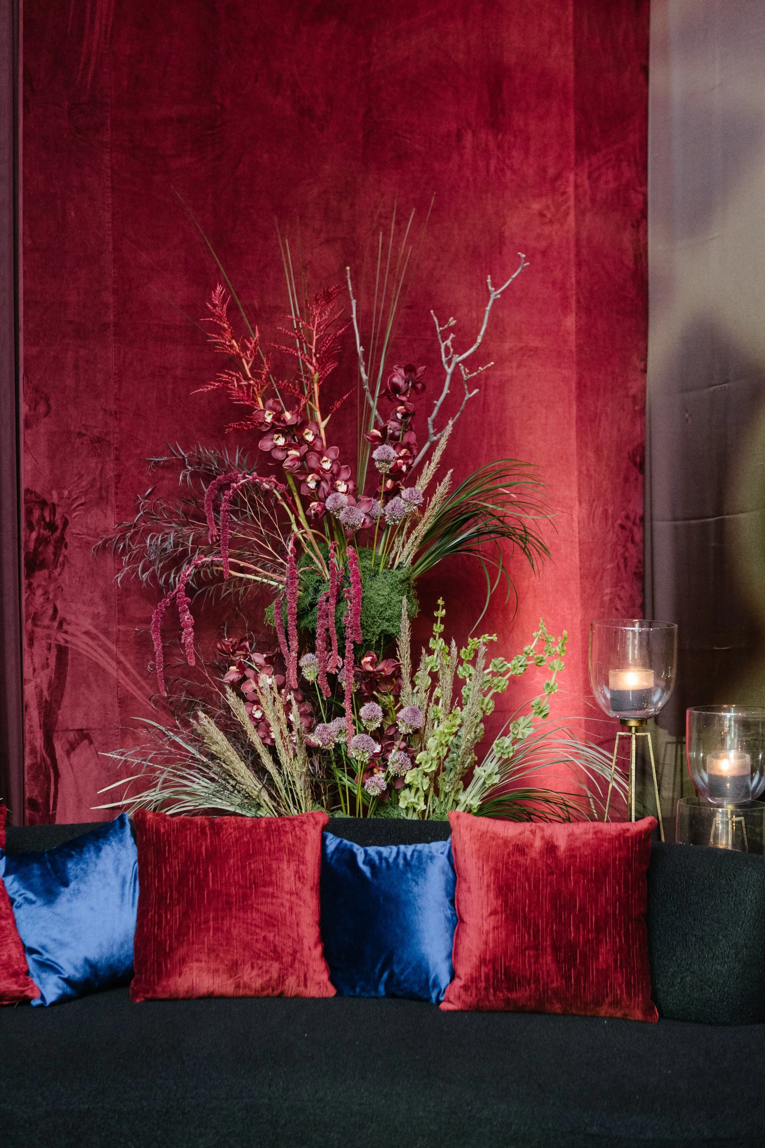 A decorative floral arrangement with various pink, purple, and green plants behind a black sofa decorated with red and blue velvet pillows, set against a red textured wall with glass candle holders.