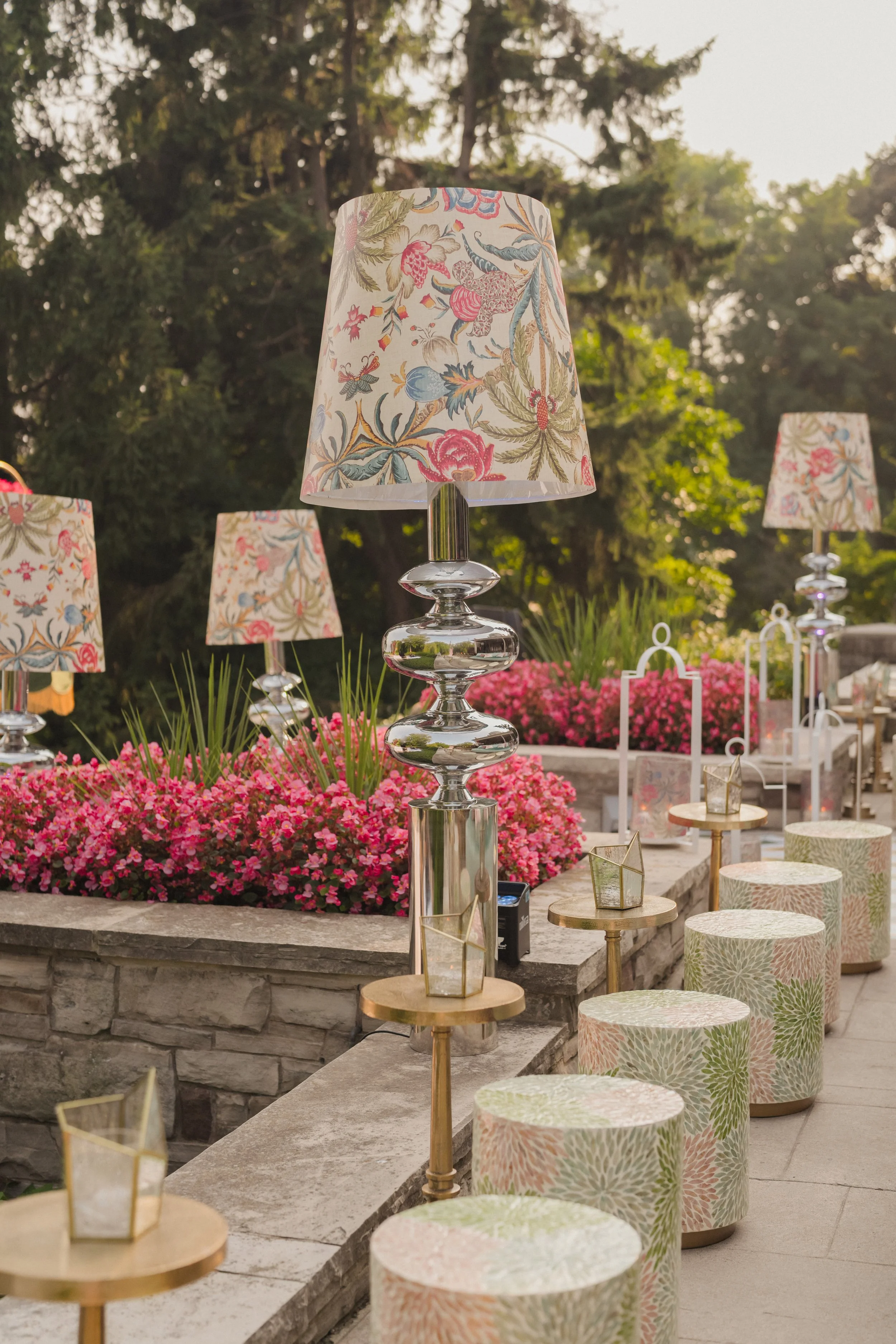 Outdoor seating area with floral lamps, small round tables, floral-patterned stools, and pink flowers in the background.