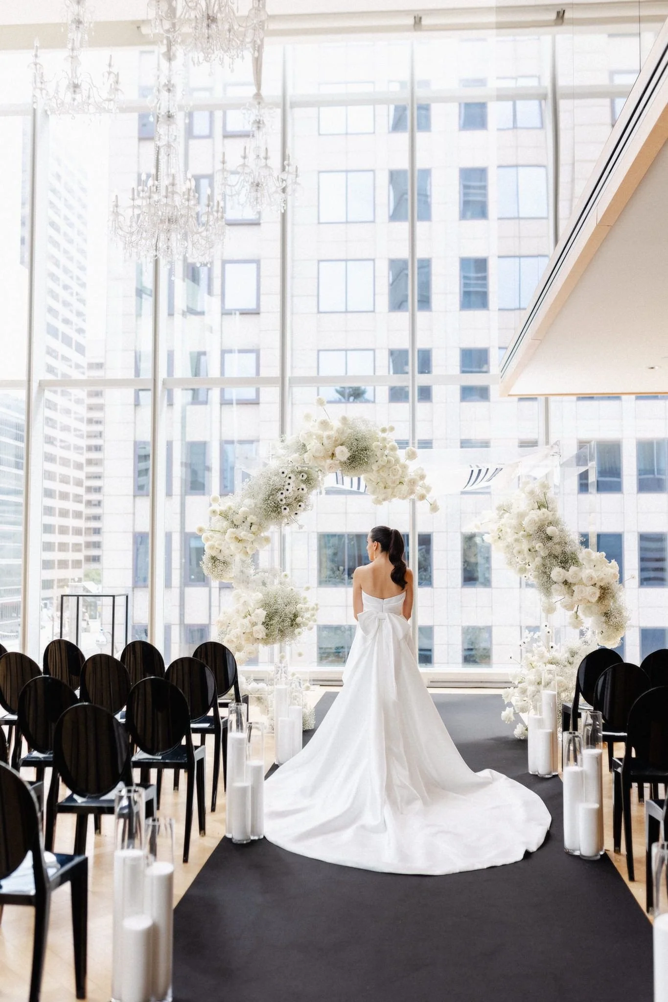 Bride in white wedding dress standing in front of a floral arch, with chairs on either side, in a modern indoor wedding venue with large windows and city buildings outside.