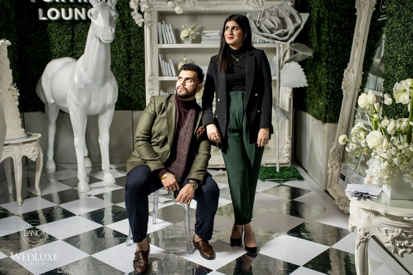 A man is sitting on a transparent chair, and a woman is standing beside him, both dressed in stylish, formal clothing. The background is decorated with white flowers, a white horse sculpture, and green foliage, creating an elegant setting.