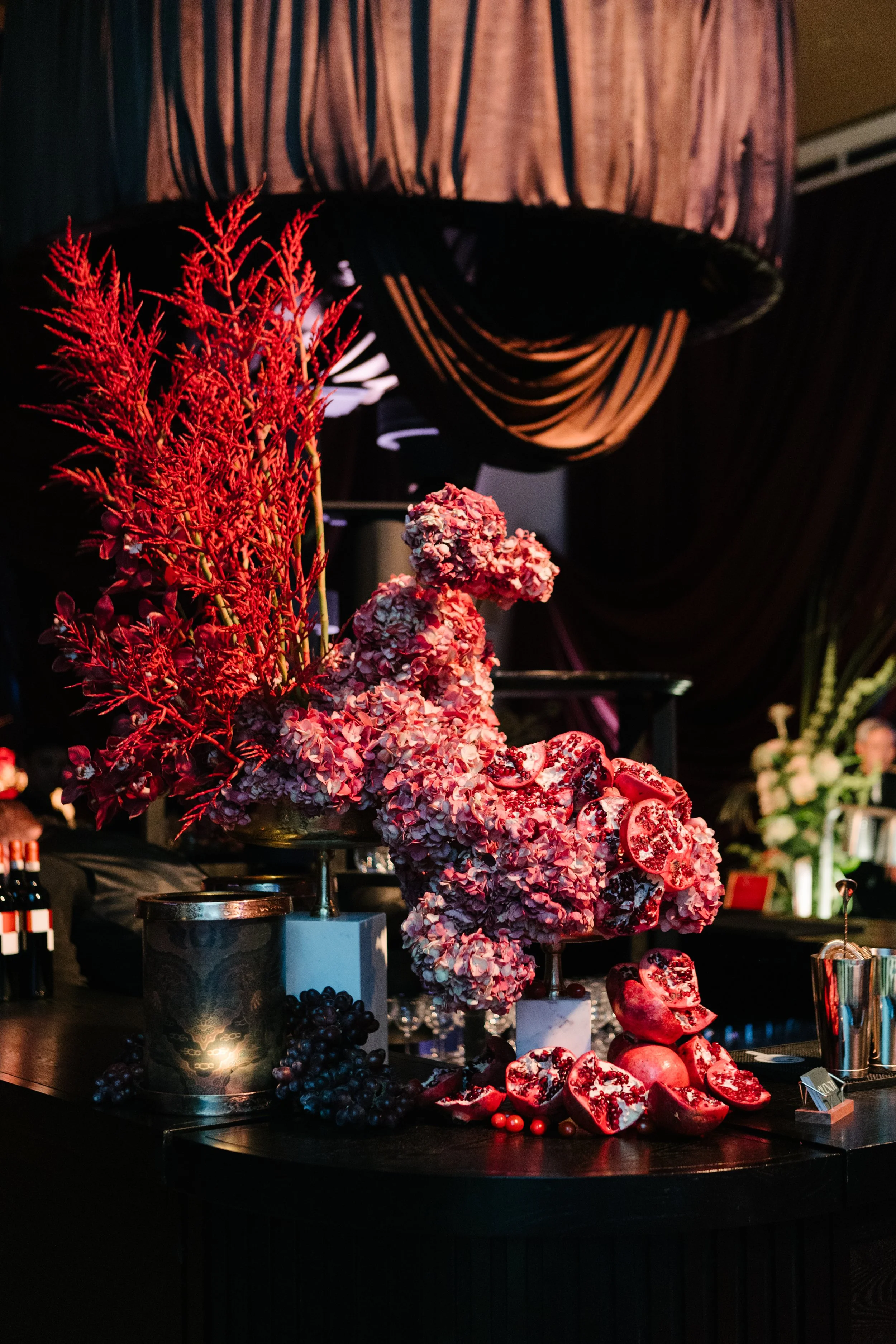 Decorative floral arrangement with pink flowers, red branches, and pomegranate halves on a black table at a formal event.