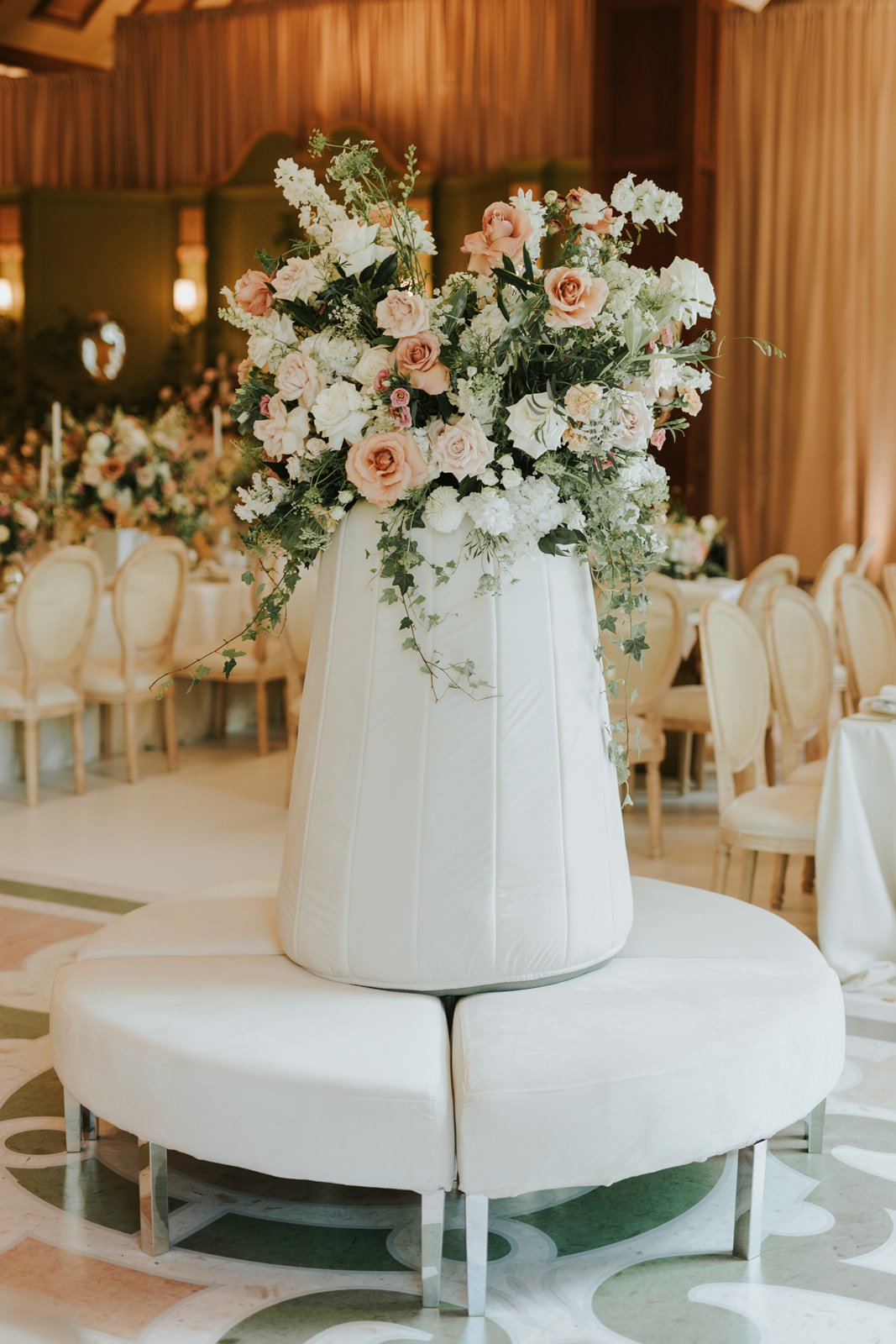 Large floral arrangement with pastels and white flowers in a white vase on a round white upholstered table in a decorated event space.