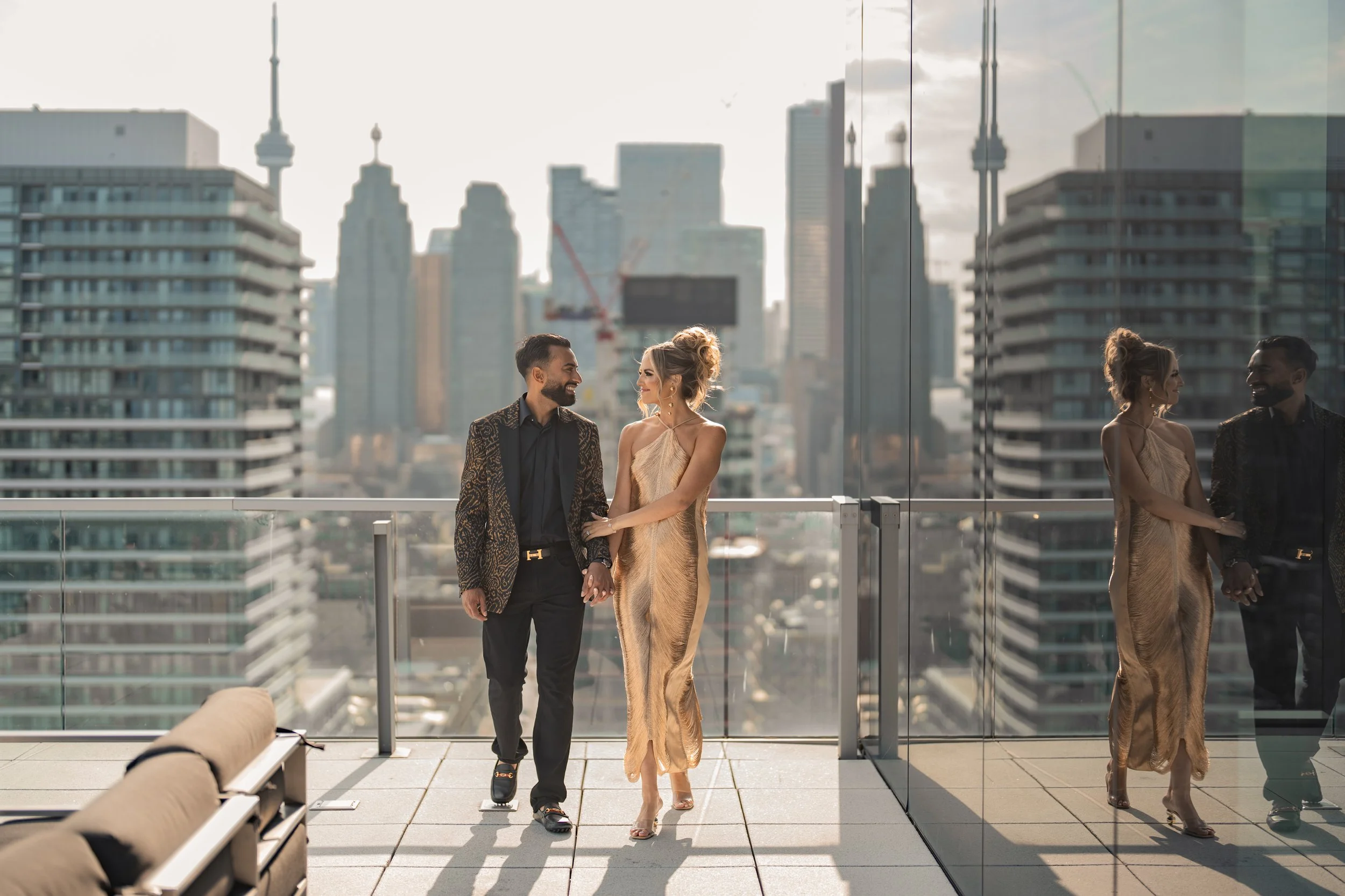 A couple holding hands and smiling on a balcony in a city with tall buildings, one in a metallic gold dress and the other in a leopard-print jacket and black pants, with their reflection visible on a glass wall.