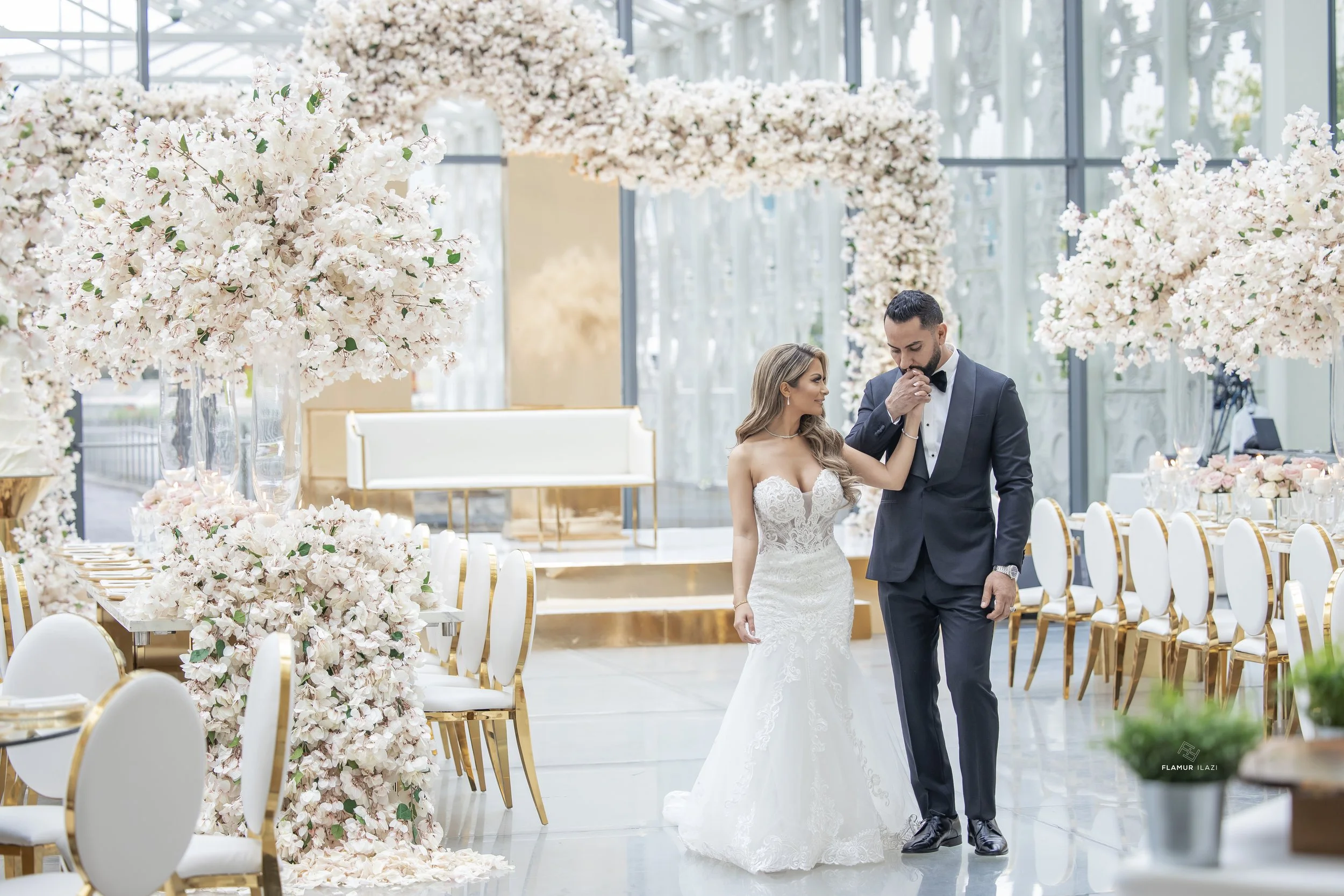 A bride and groom sharing a kiss at their wedding reception in a decorated indoor venue with large floral arrangements and elegant chairs.