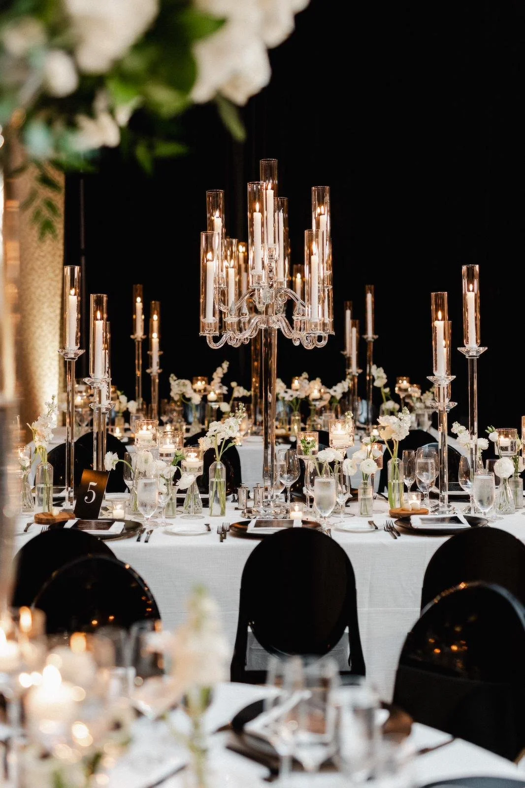 Elegant banquet table set for a formal event with a large crystal chandelier centerpiece, white flowers in glass vases, black chairs, and candlelit decor.