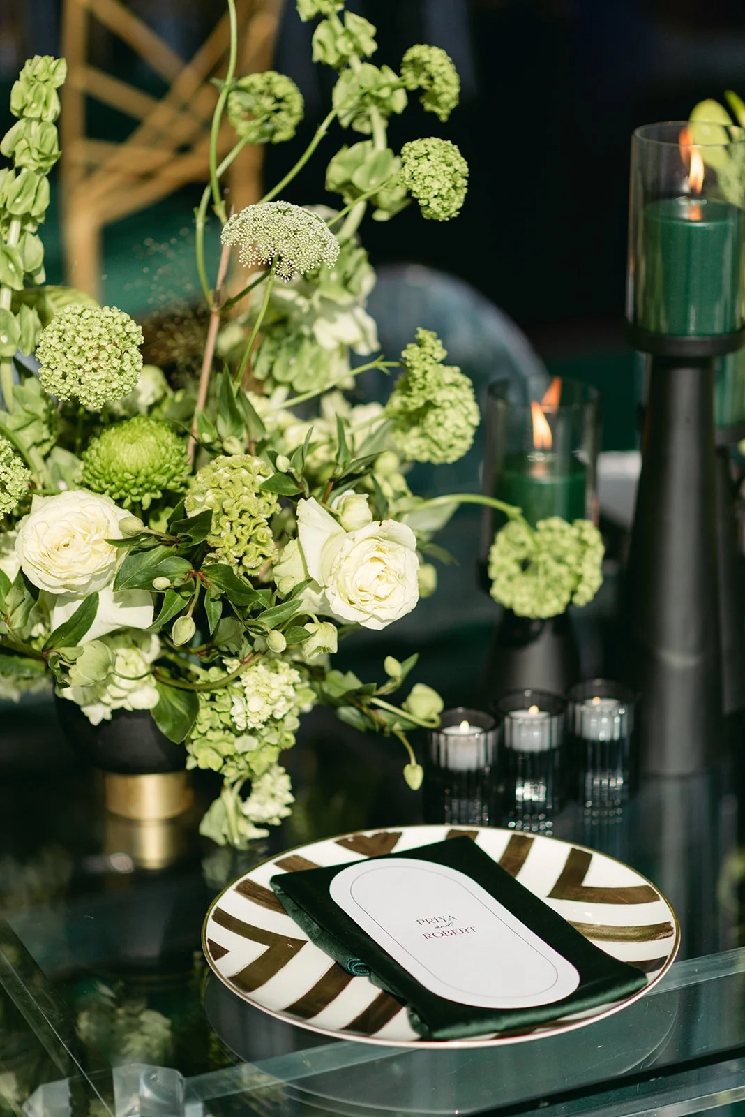 Elegant table setting with a black and white striped plate, a folded black napkin, and a card that reads 'PRIVA LOVE ROBERT.' A large floral arrangement of white roses, green hydrangeas, and green blooms, along with tall green candles and small votiv