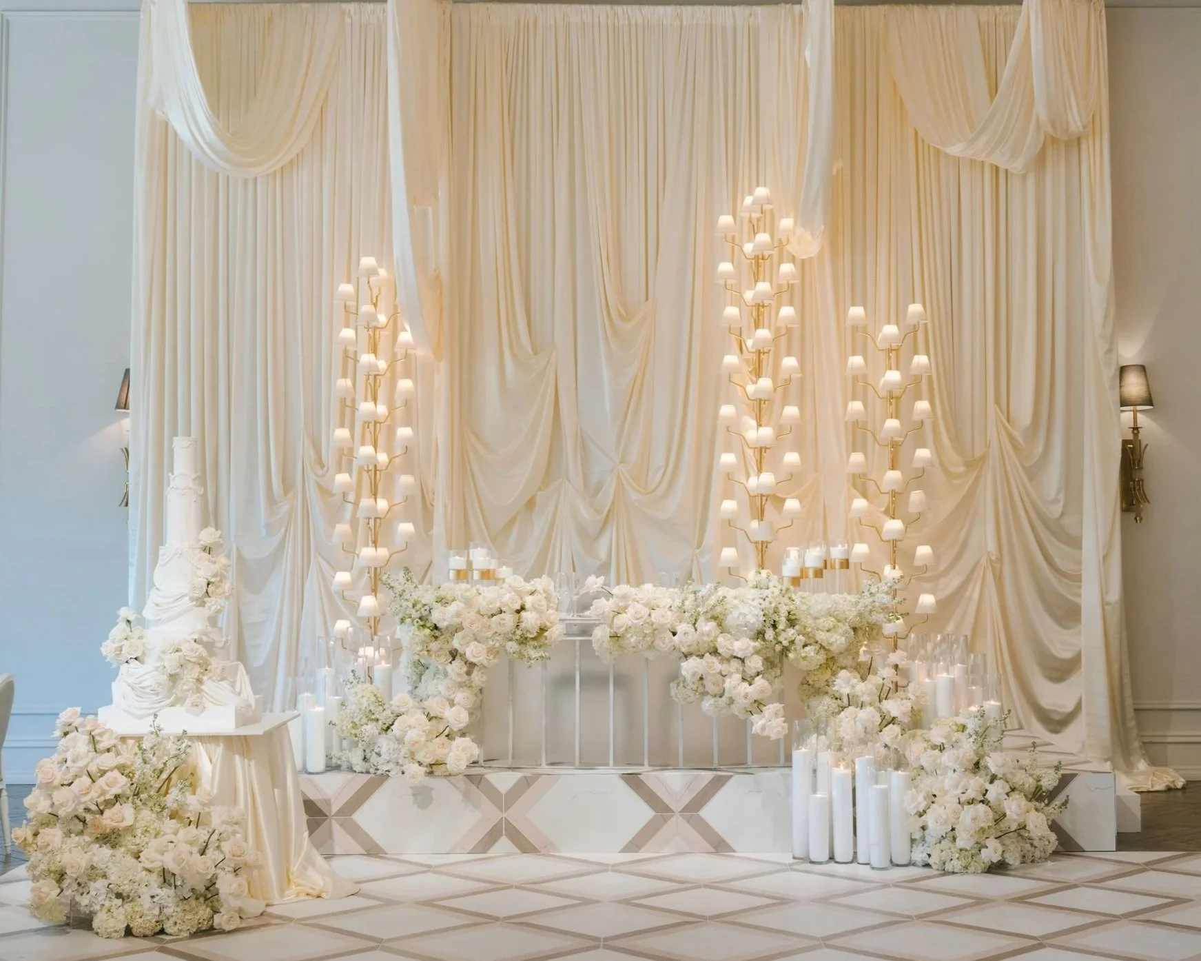 Elegant wedding or event backdrop with cream-colored drapery, chandeliers, and floral arrangements of white flowers, with candles and candles on the floor.