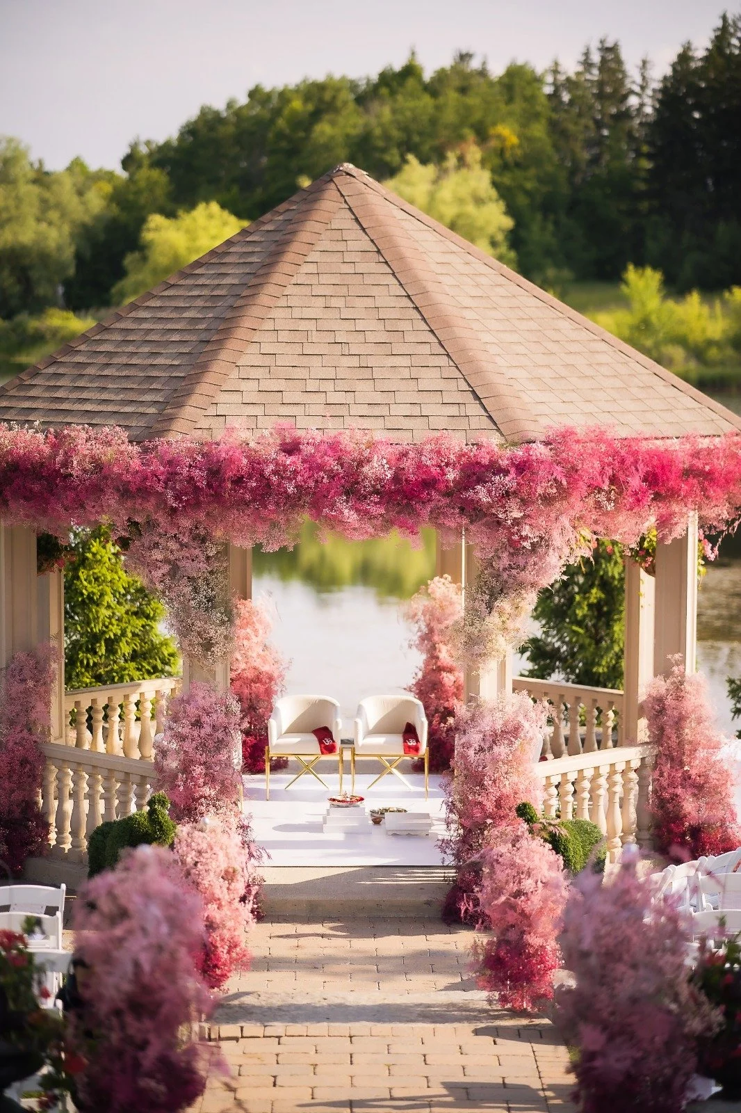 A decorated outdoor wedding gazebo covered with pink and white flowers, with a pathway leading to white chairs, set against a background of green trees.