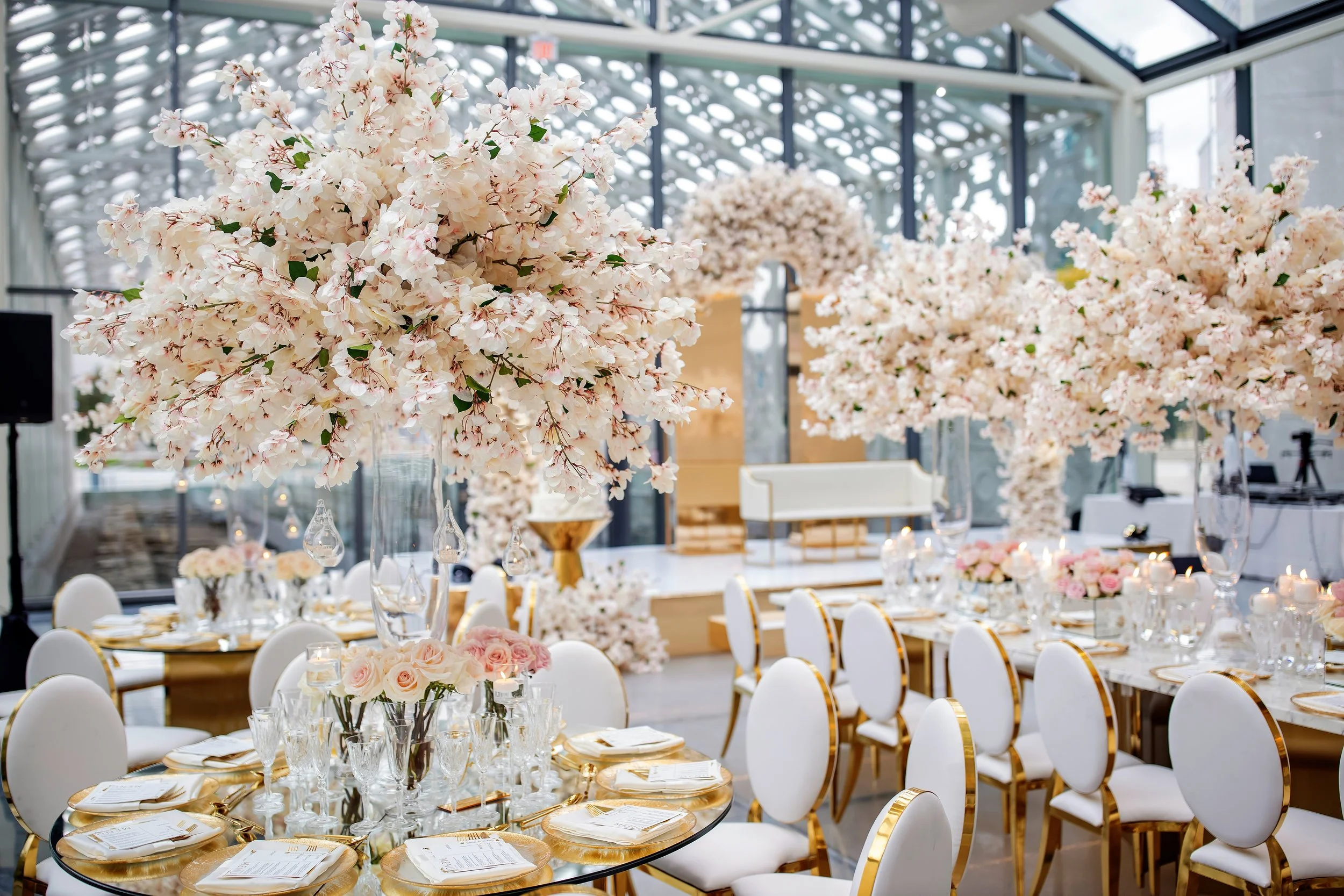 Elegant event space with large pink cherry blossom trees, round tables with gold trim, white chairs, floral centerpieces, and soft lighting.