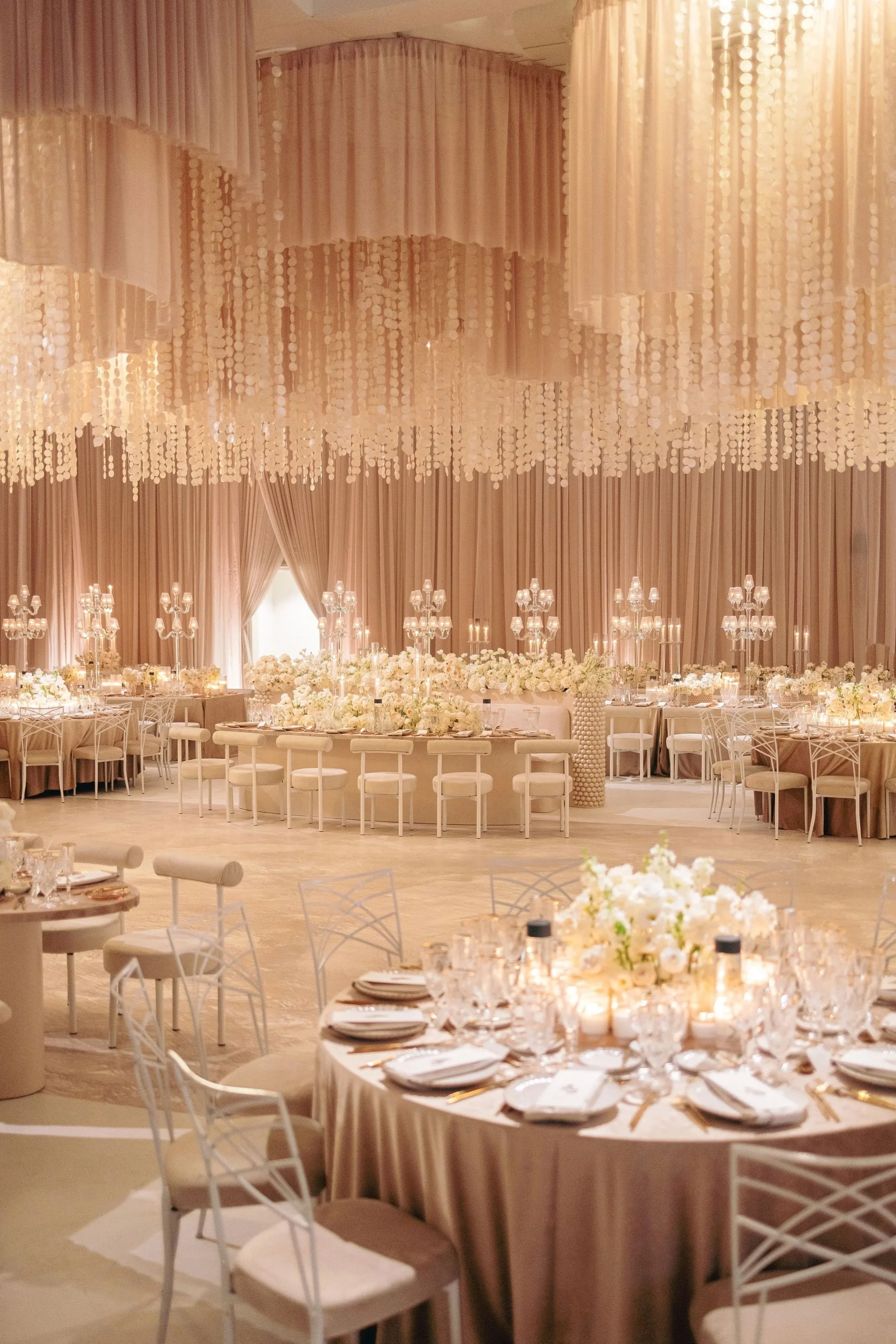 Elegant banquet hall decorated for a wedding reception with pastel pink draped curtains, hanging crystal accents, numerous floral centerpieces, and candlelit tables.