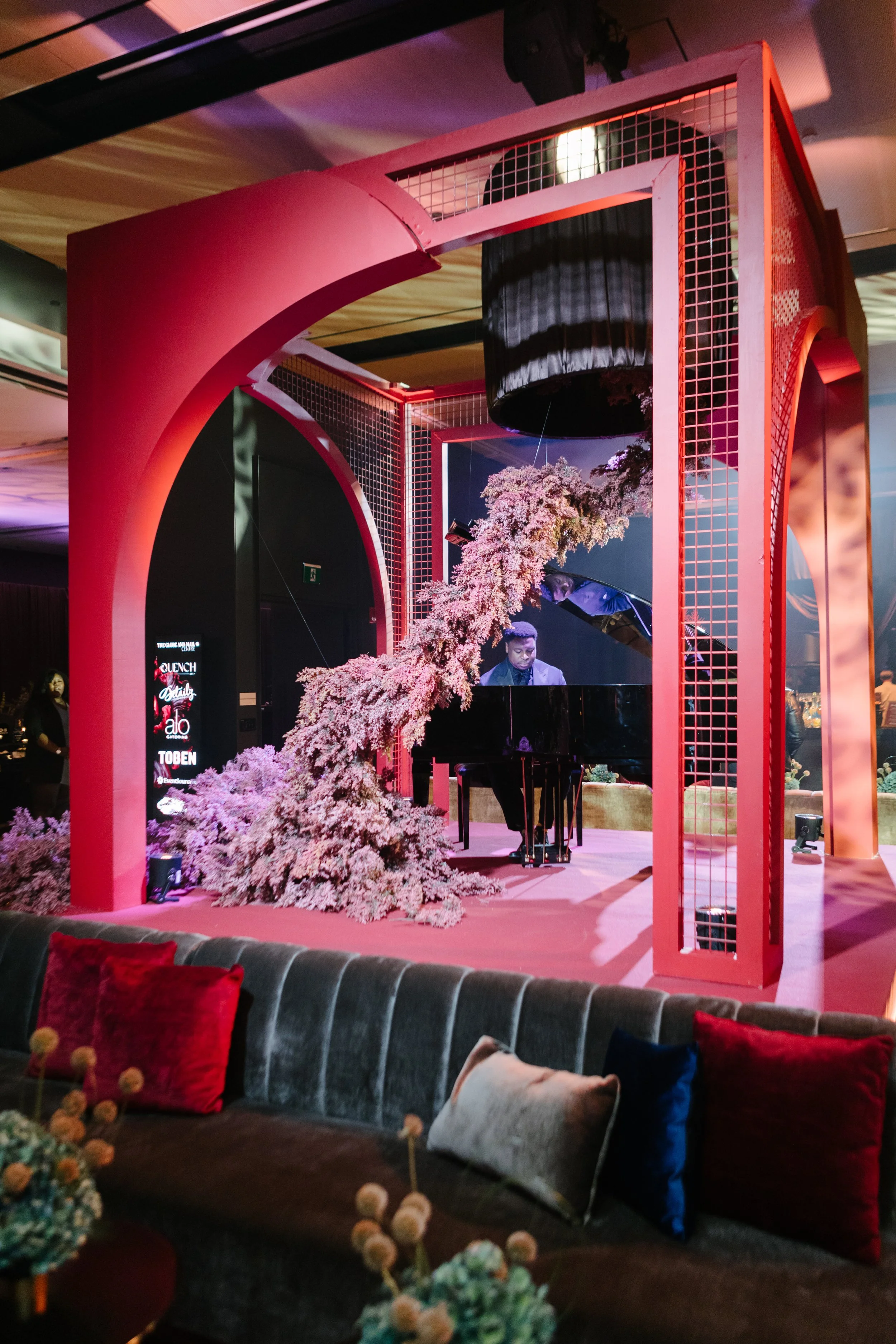A person playing a grand piano on a decorated stage with pink floral arrangements and modern red architecture, in a dimly lit room with seating and decorative pillows.
