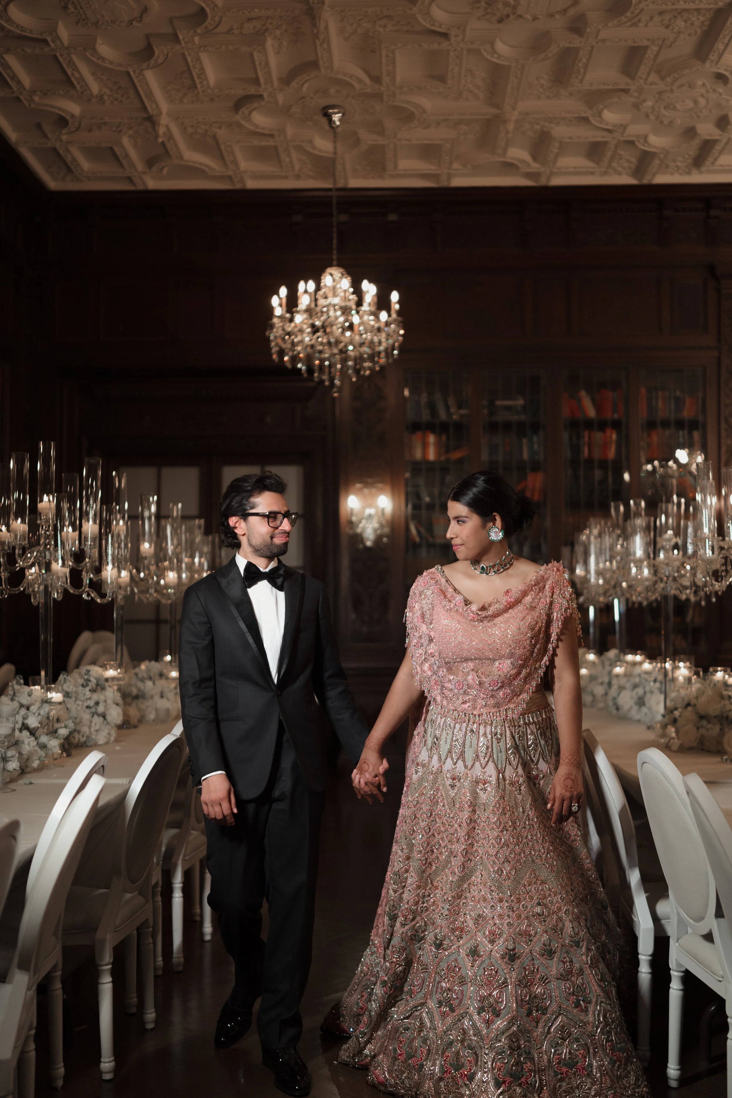 A couple dressed in formal attire walking hand in hand in an elegant, dimly lit dining room with ornate ceiling, chandeliers, and a long table decorated with white flowers and candelabras.
